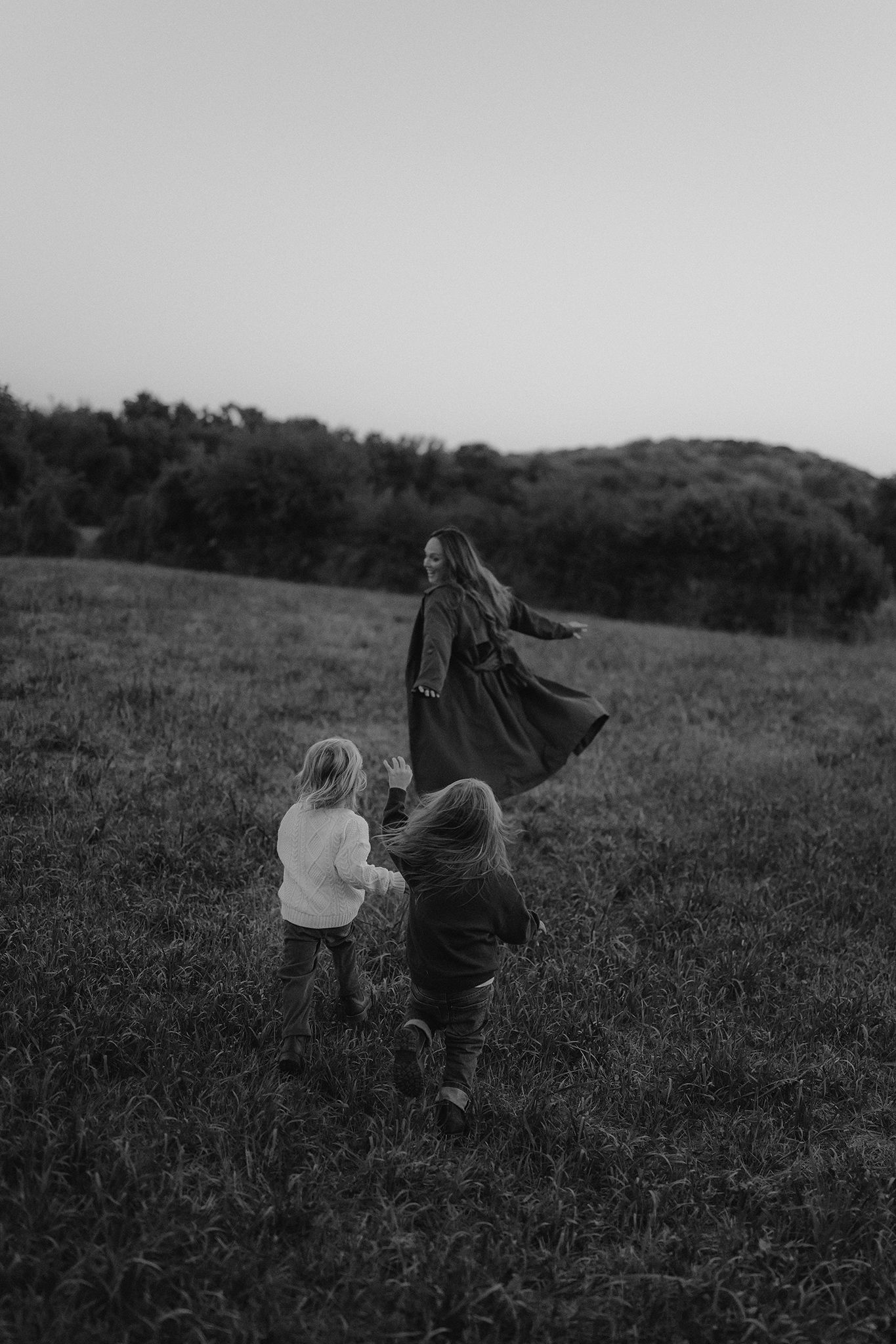Black and white photo of a mother in a flowing dress playing with two young children in a grassy field at dusk.