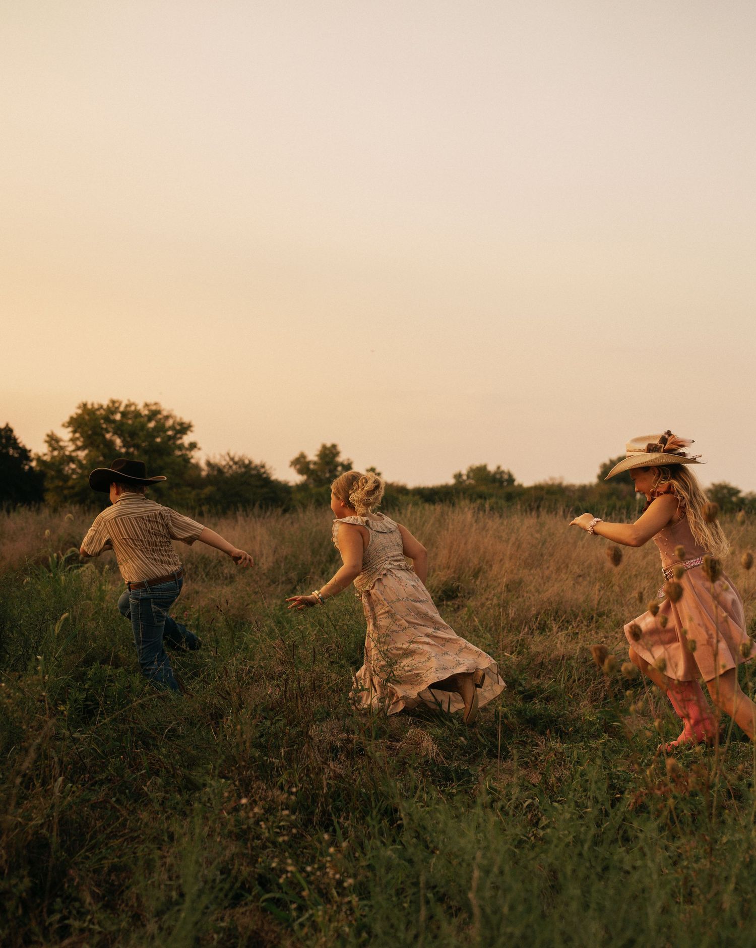 Three children run through a golden field at sunset wearing floral dresses and a cowboy hat.