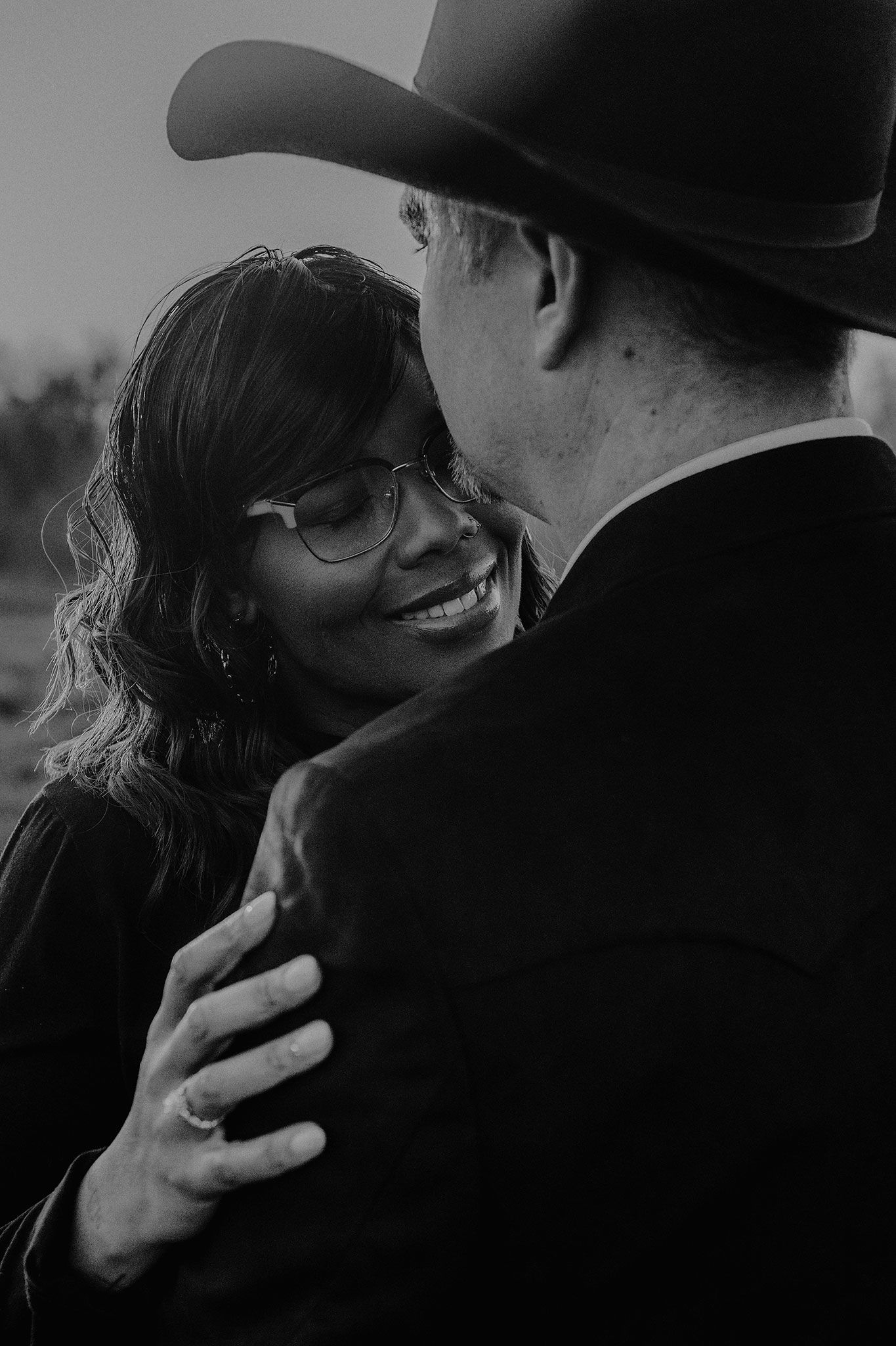 Black and white photo of a smiling woman with glasses embracing a man in a cowboy hat.