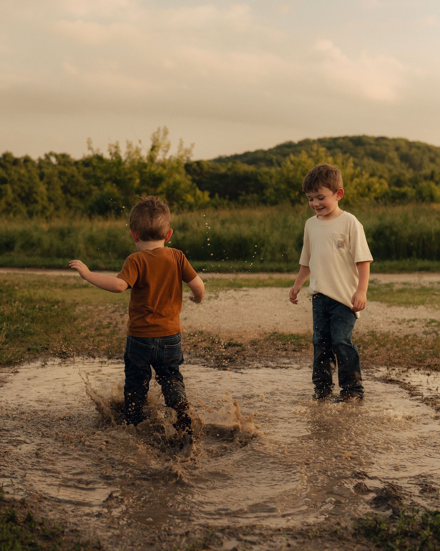 Two young boys splashing joyfully in a muddy puddle outdoors, wearing casual clothes with green hills in the background.