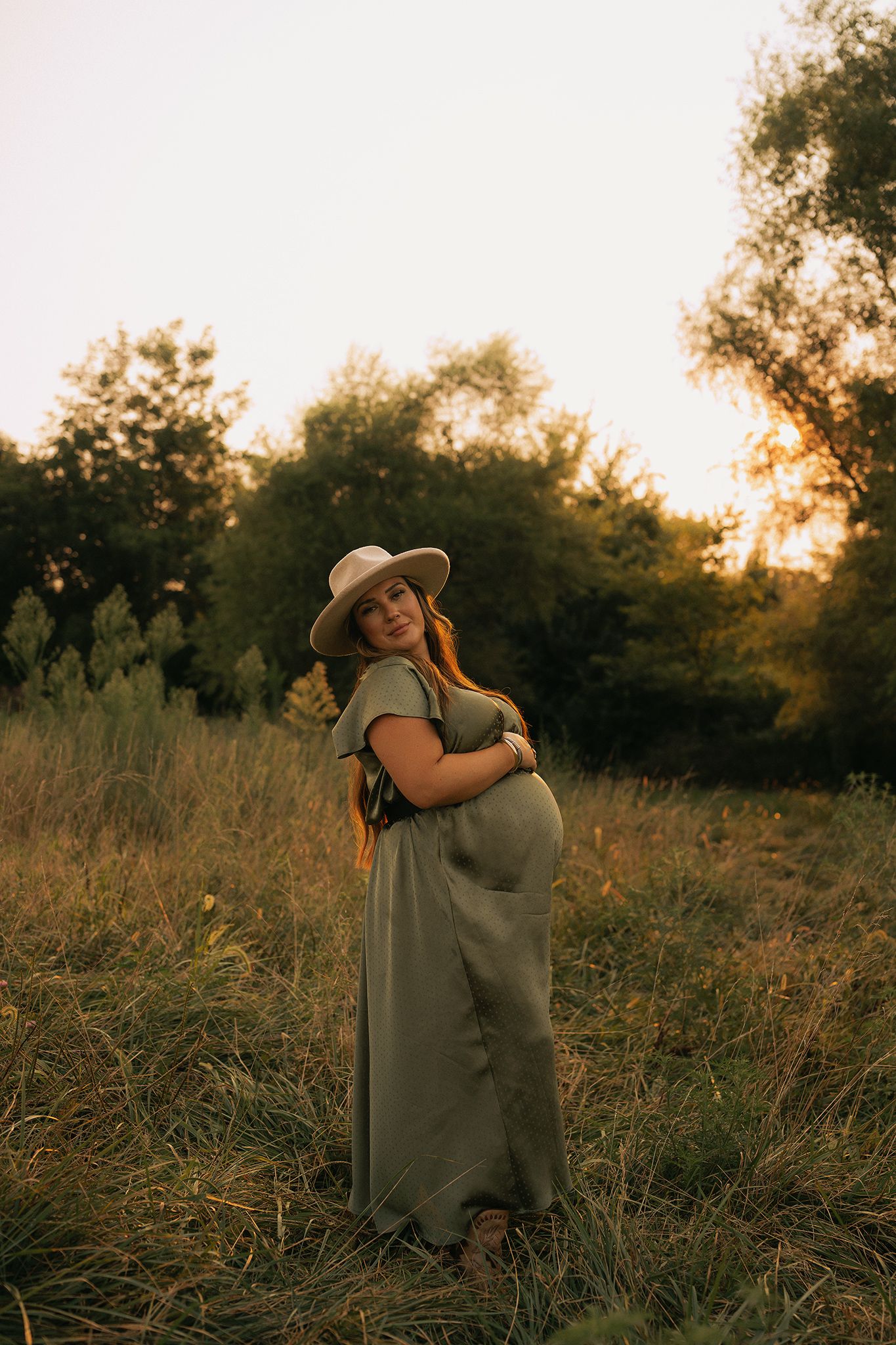 Pregnant woman in olive maxi dress and straw hat cradling baby bump in golden sunset field.