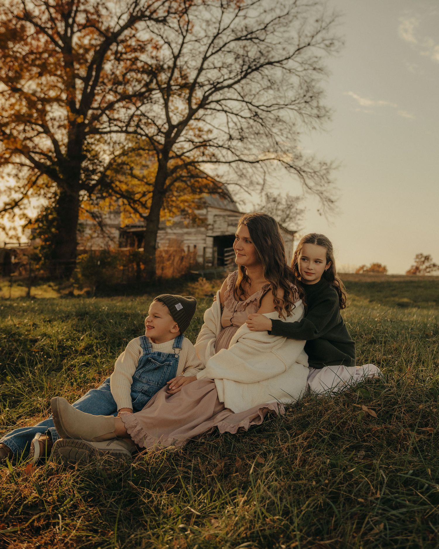A mother sits on grass with two young daughters at golden hour, with an old farmhouse and autumn trees in the background.