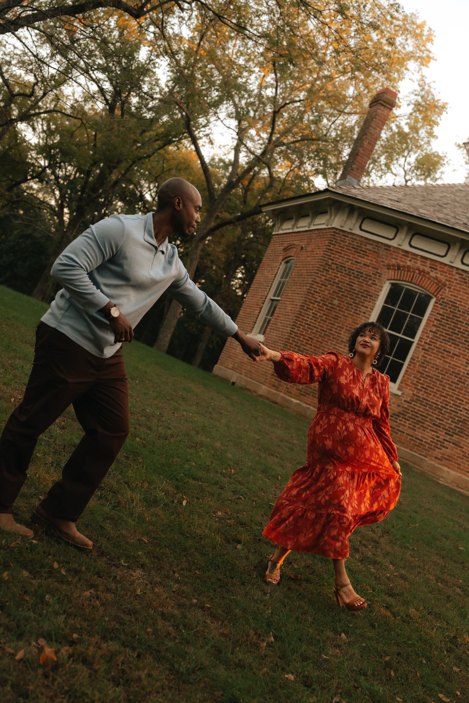 A couple dances joyfully on a green lawn outside a brick building surrounded by autumn trees.