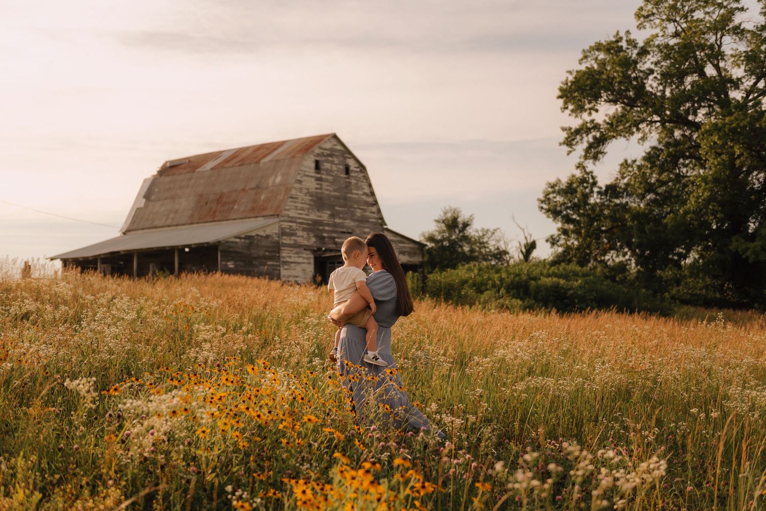 A mother holds her child in a wildflower field with a rustic barn and tree at golden hour.