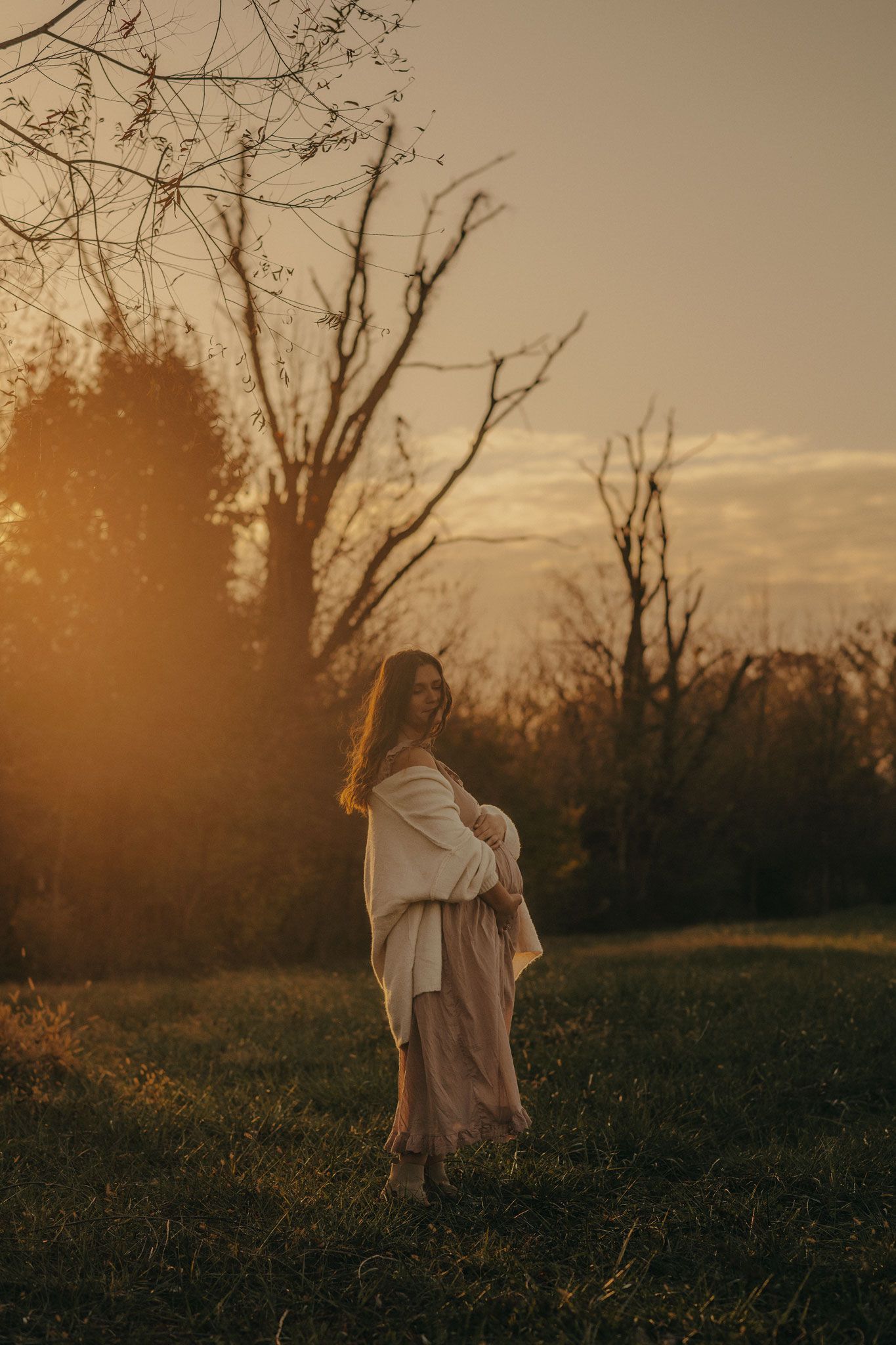 A woman in a white top and pink skirt stands in a golden sunset field surrounded by bare trees.
