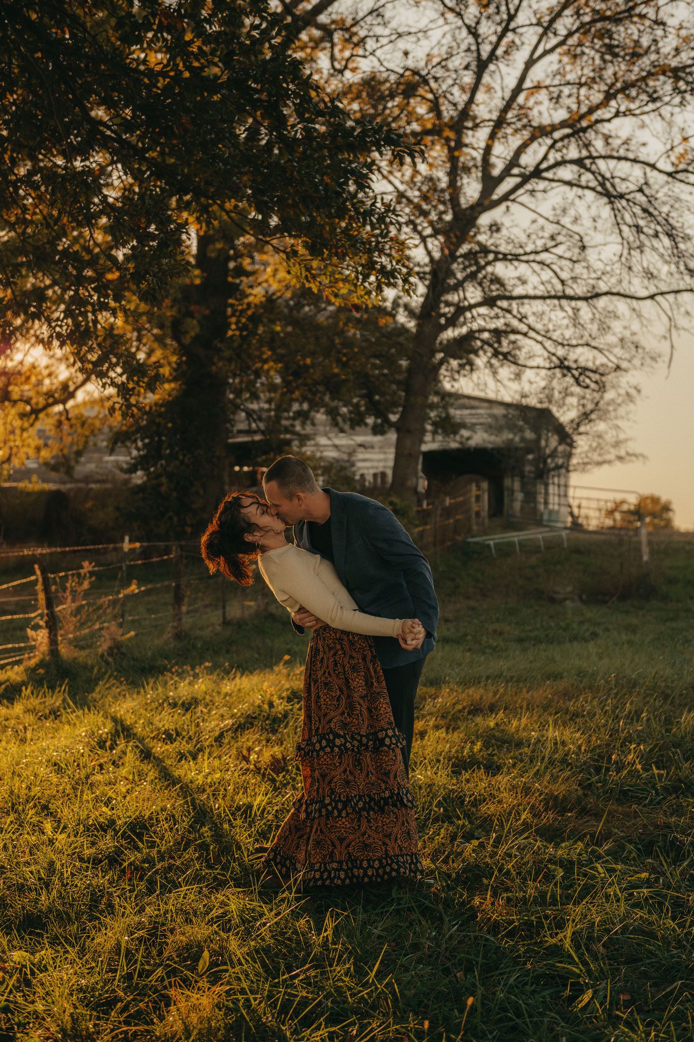 Couple sharing a romantic kiss at golden hour in a lush green field with trees and a rustic barn backdrop.
