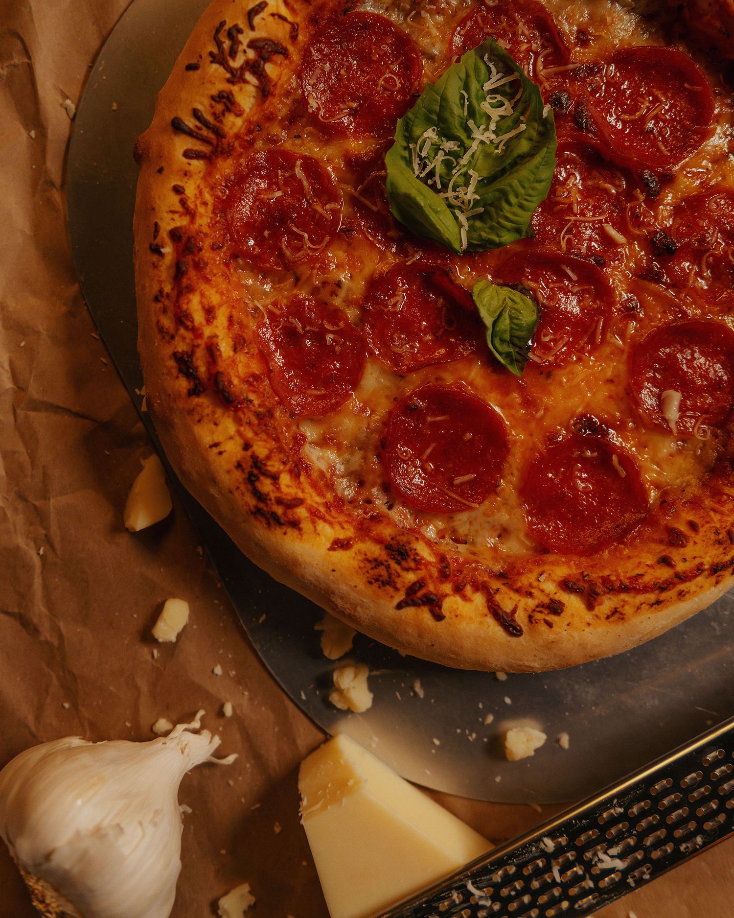 Pepperoni pizza topped with fresh basil and shredded cheese on a pan, with garlic and parmesan nearby.
