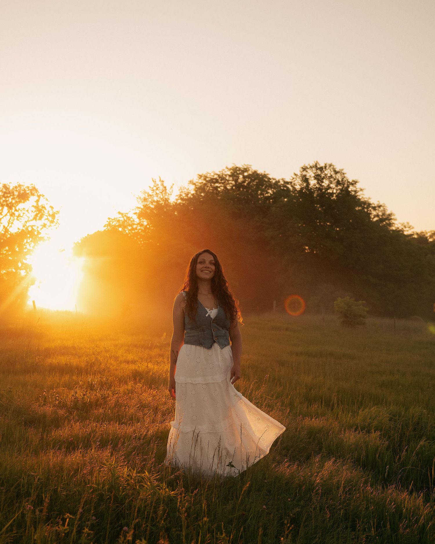 Woman in white dress and denim vest standing in golden sunset field with trees in background.