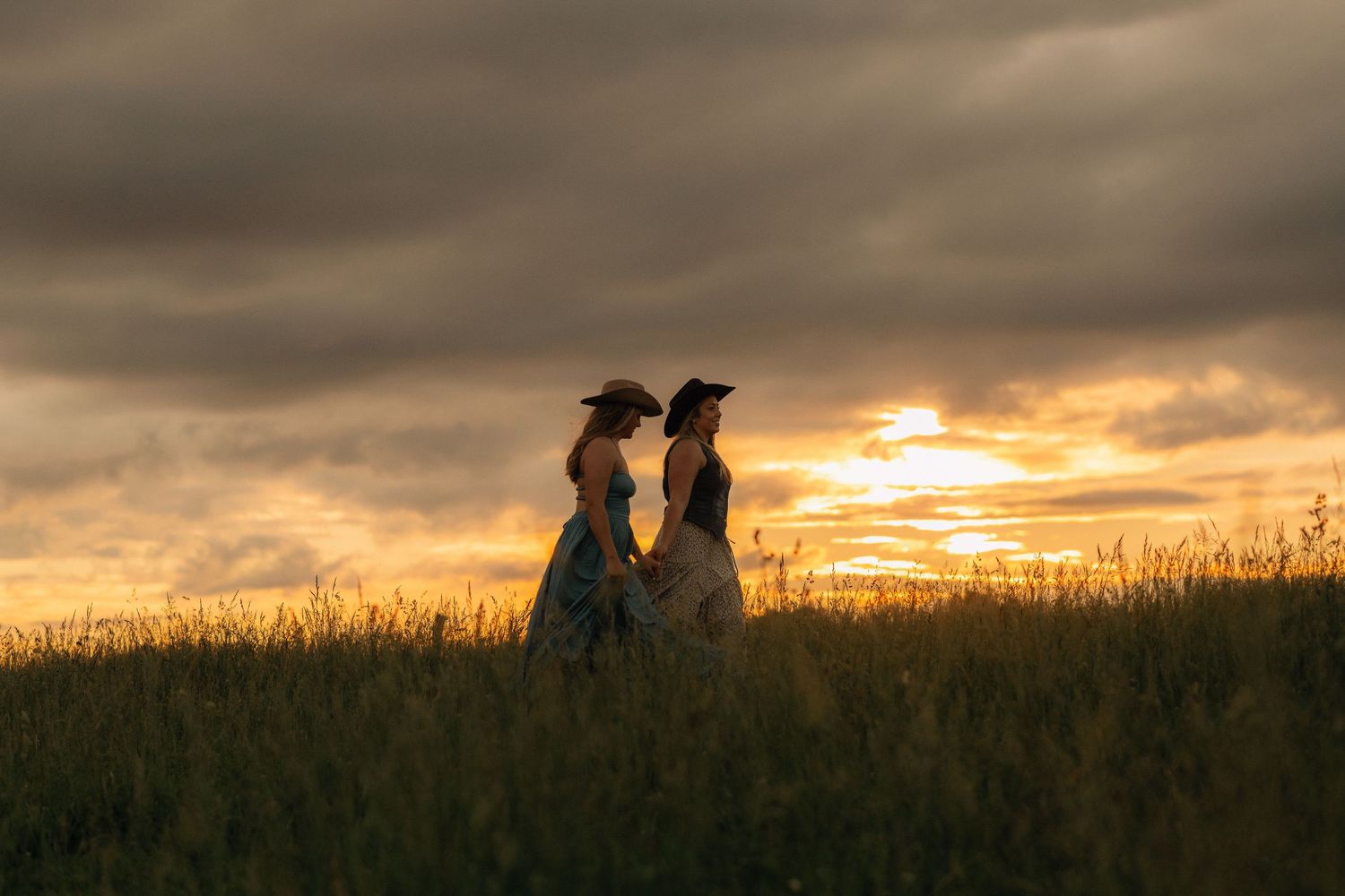 Two women in hats and dresses stand in a golden field at sunset under dramatic cloudy skies.