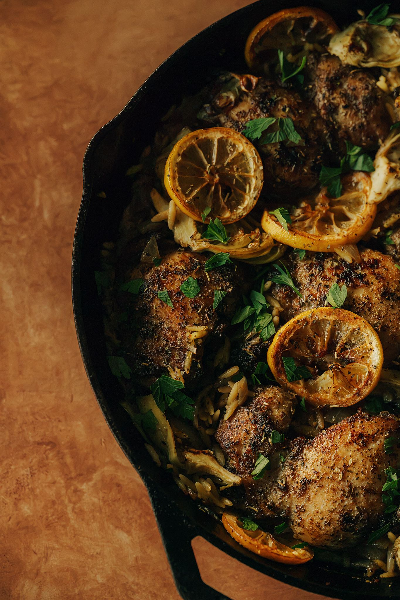 Cast iron skillet with roasted chicken thighs, caramelized lemon slices, fennel, and fresh herbs on rustic background.