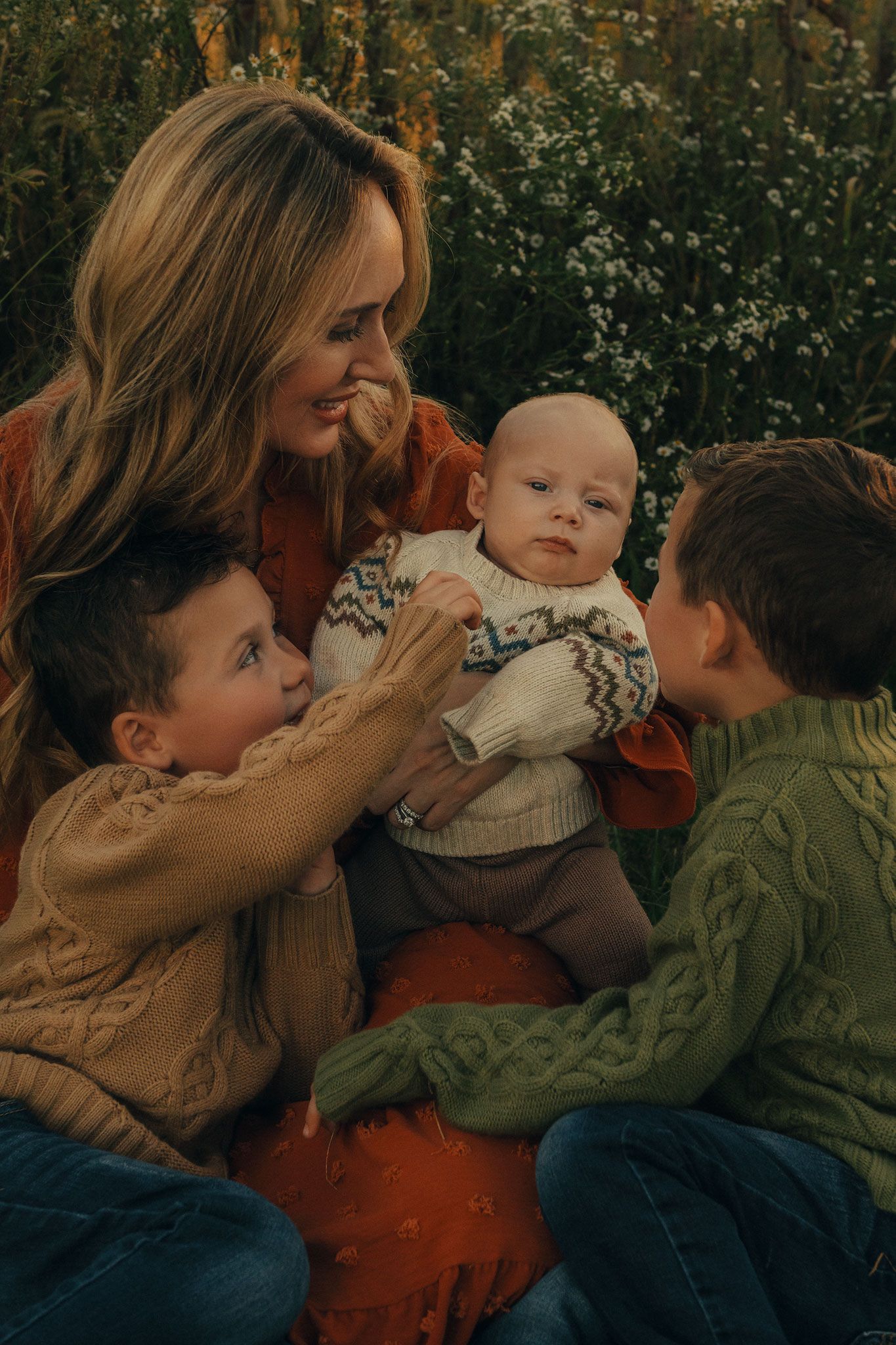 A smiling mother sits outdoors with her three young sons, all wearing cozy sweaters, sharing a warm family moment.