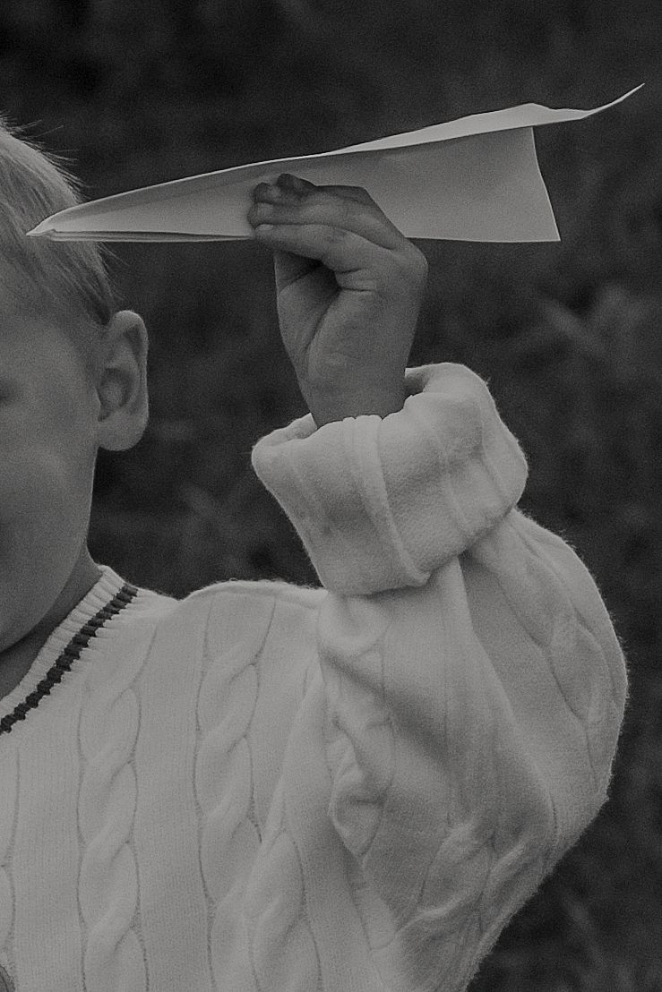 Black and white photo of a child in a cable-knit sweater holding a paper airplane ready to throw it.