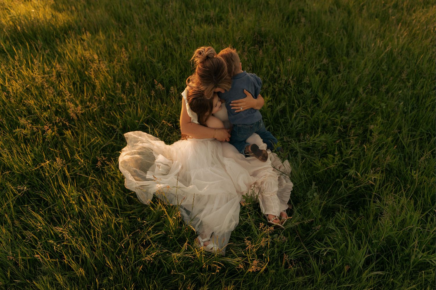 A mother in a white dress embraces her young child in a sunlit green meadow during golden hour.