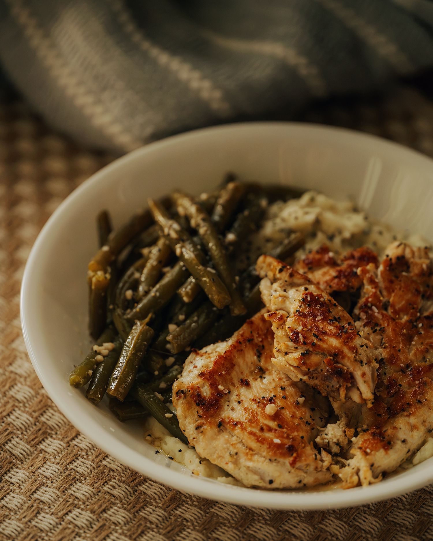 A white bowl filled with seasoned grilled chicken, green beans, and creamy mashed potatoes on a woven mat.