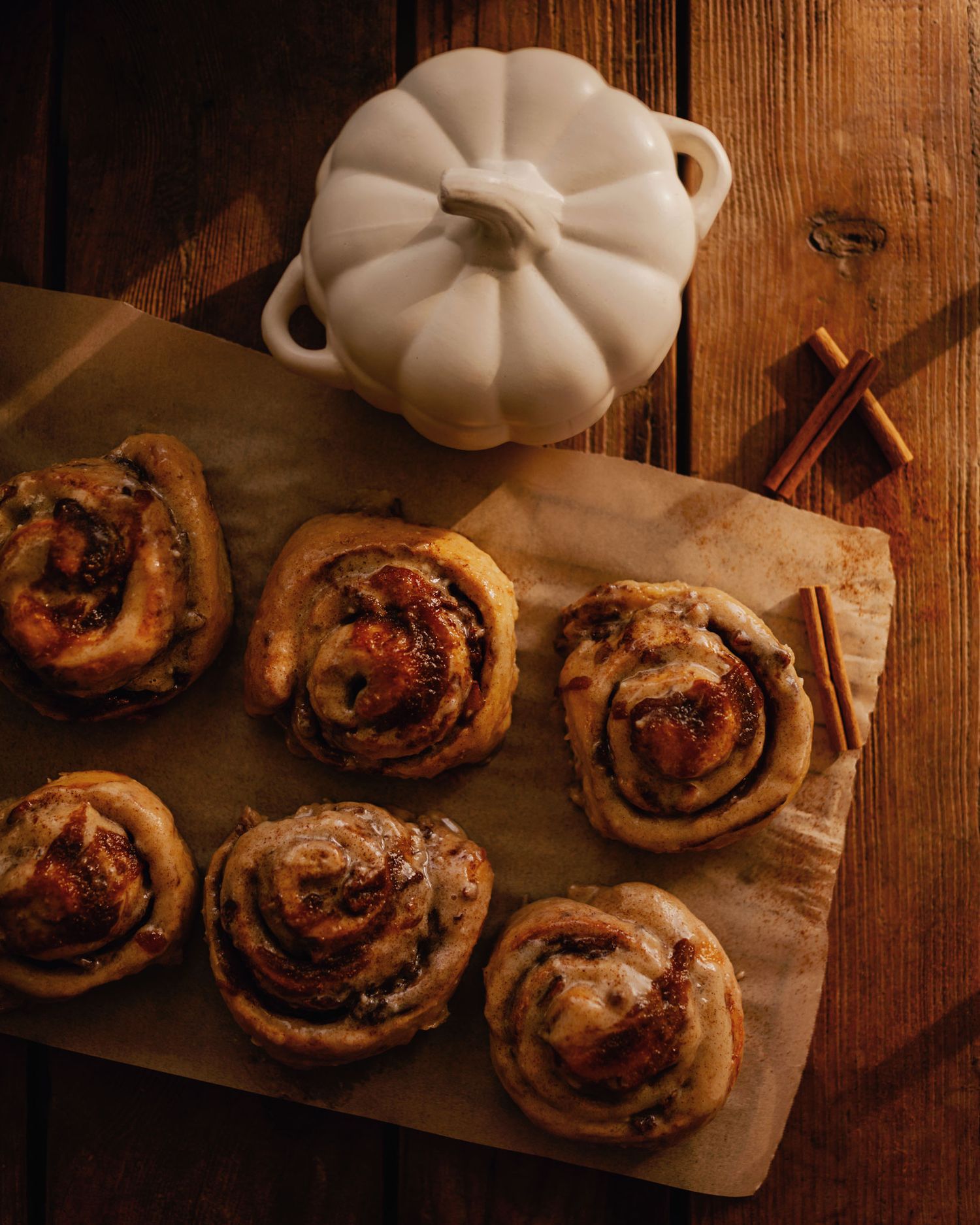 Freshly baked apple cinnamon rolls on parchment paper beside a white pumpkin dish and cinnamon sticks on a wooden board.