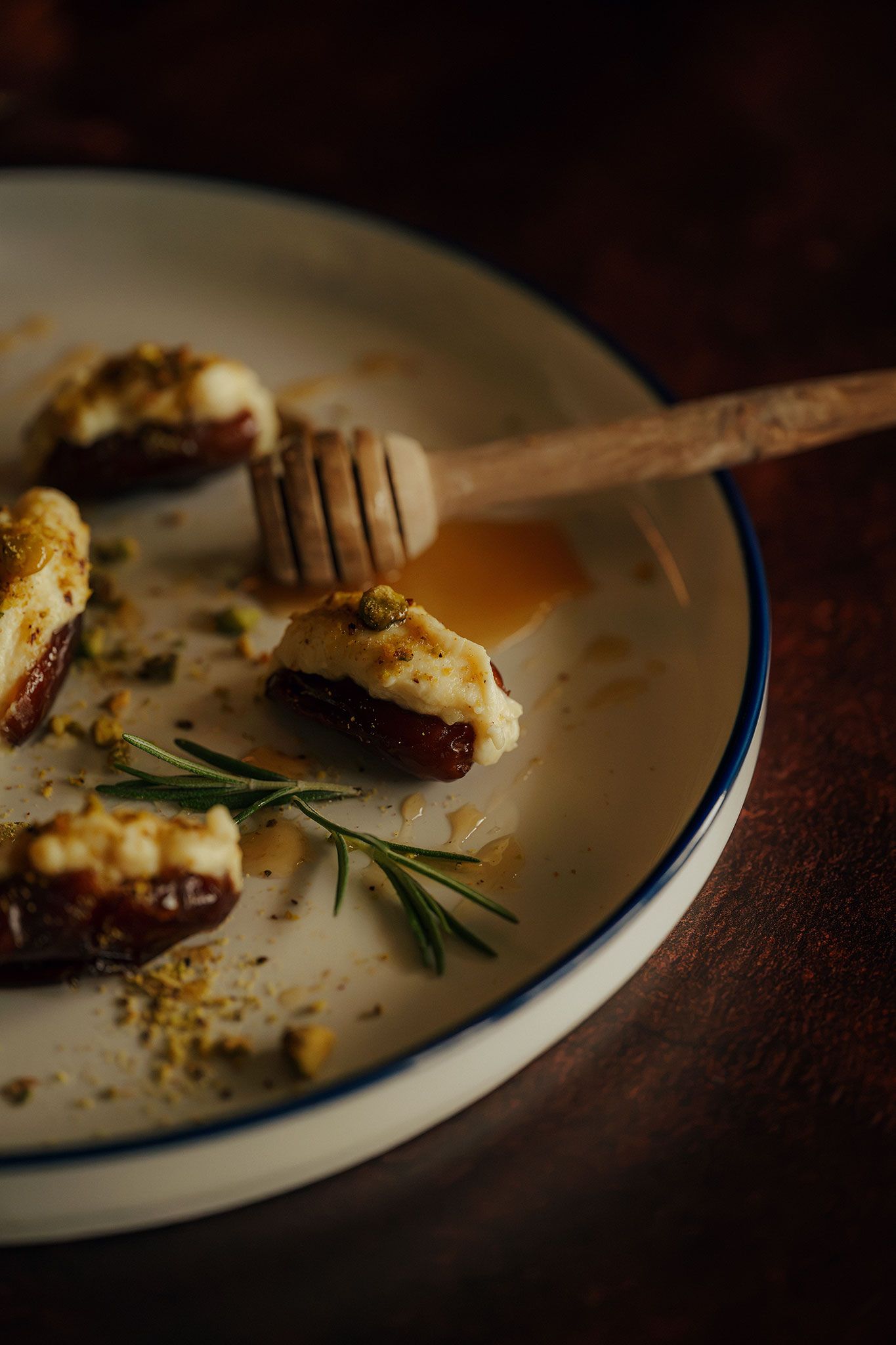 Dates stuffed with cream cheese, drizzled with honey and garnished with rosemary and pistachios on a white plate.