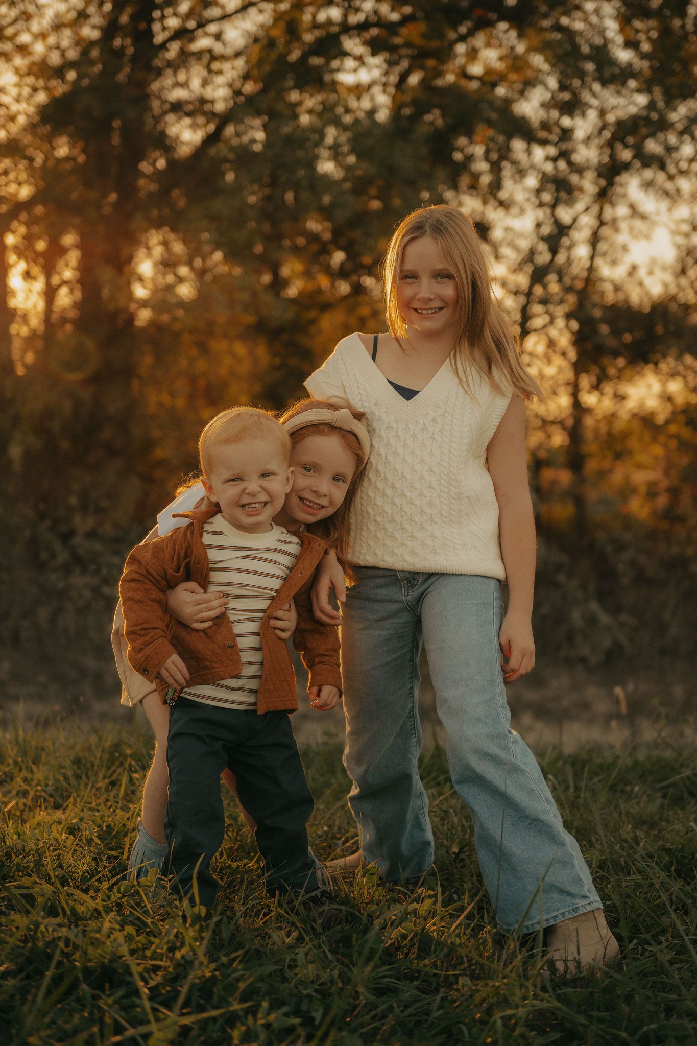 Three siblings pose together smiling in a sunlit autumn field at golden hour.