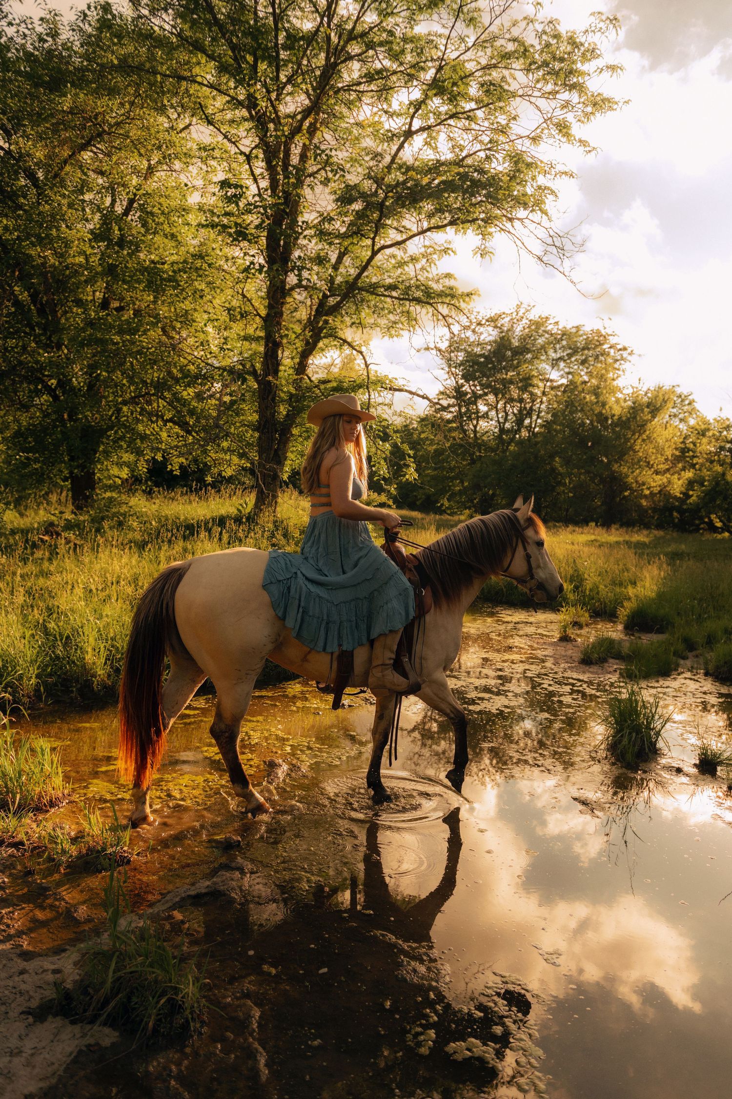 Woman in blue dress and straw hat riding a horse through a shallow stream in a lush green forest.