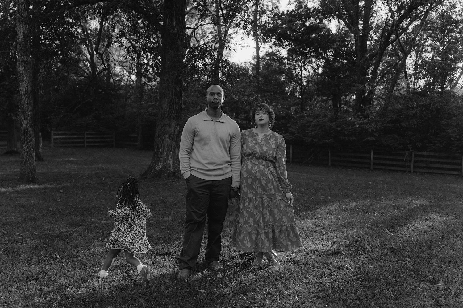 Black and white photo of a family with a young child standing on a grassy lawn surrounded by trees.