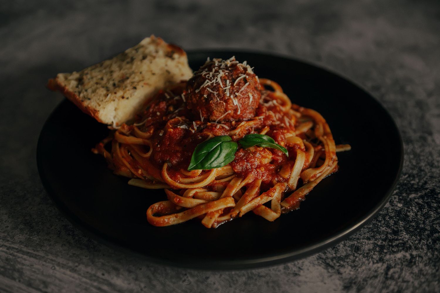 A black plate of spaghetti with meatballs in marinara sauce, fresh basil, parmesan, and garlic bread.