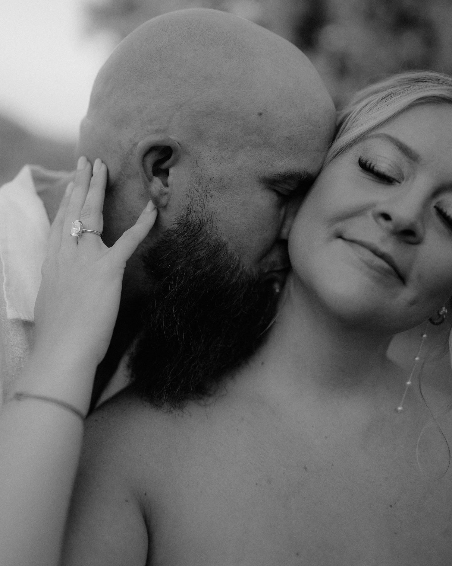 Black and white romantic couple photo with bald bearded man kissing womans cheek as she smiles wearing diamond ring.
