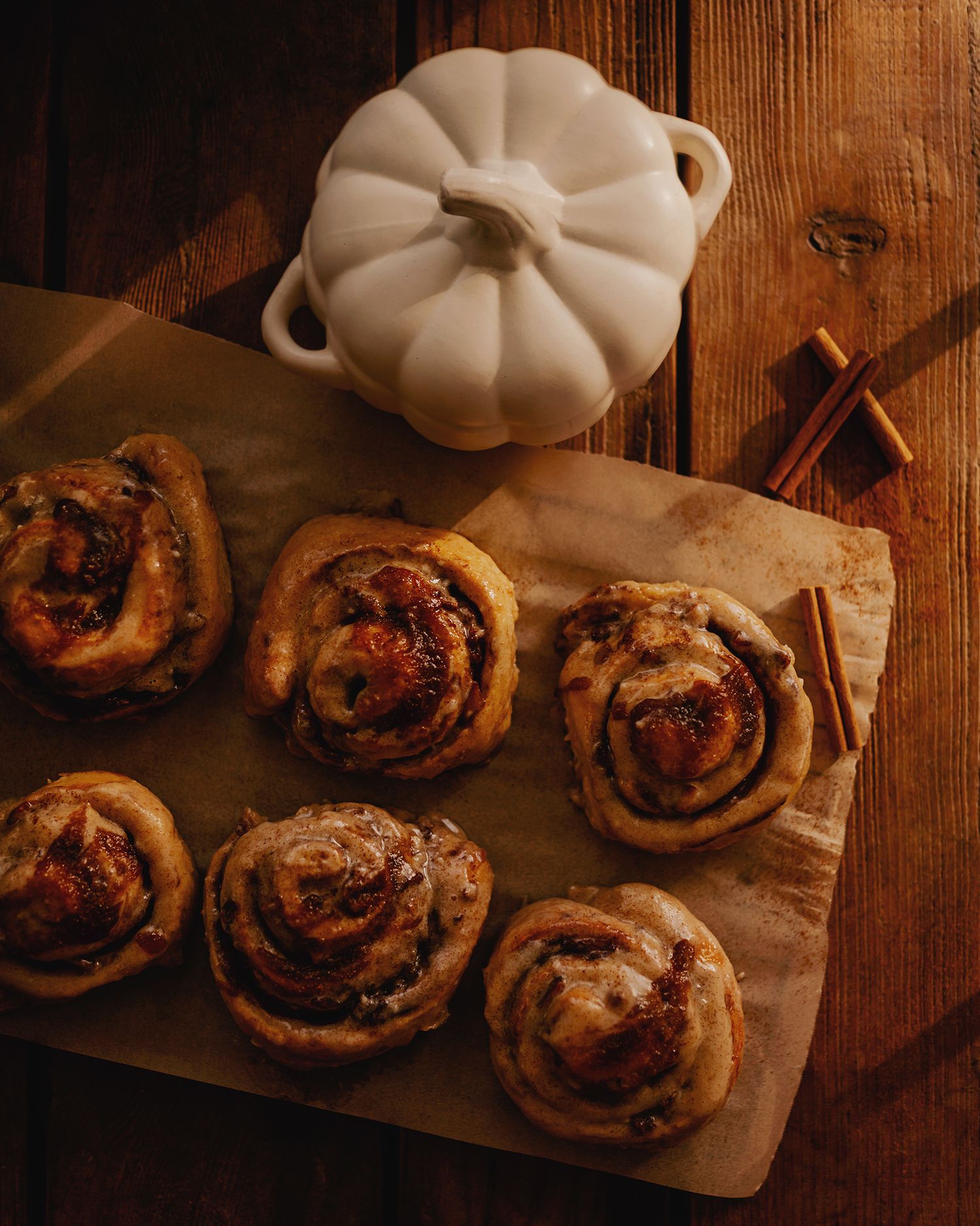 Freshly baked cinnamon rolls on parchment paper beside cinnamon sticks and a white pumpkin dish on a wooden board.