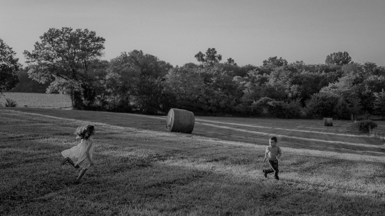 Two children run through a vast green field with a hay bale at golden hour in black and white.