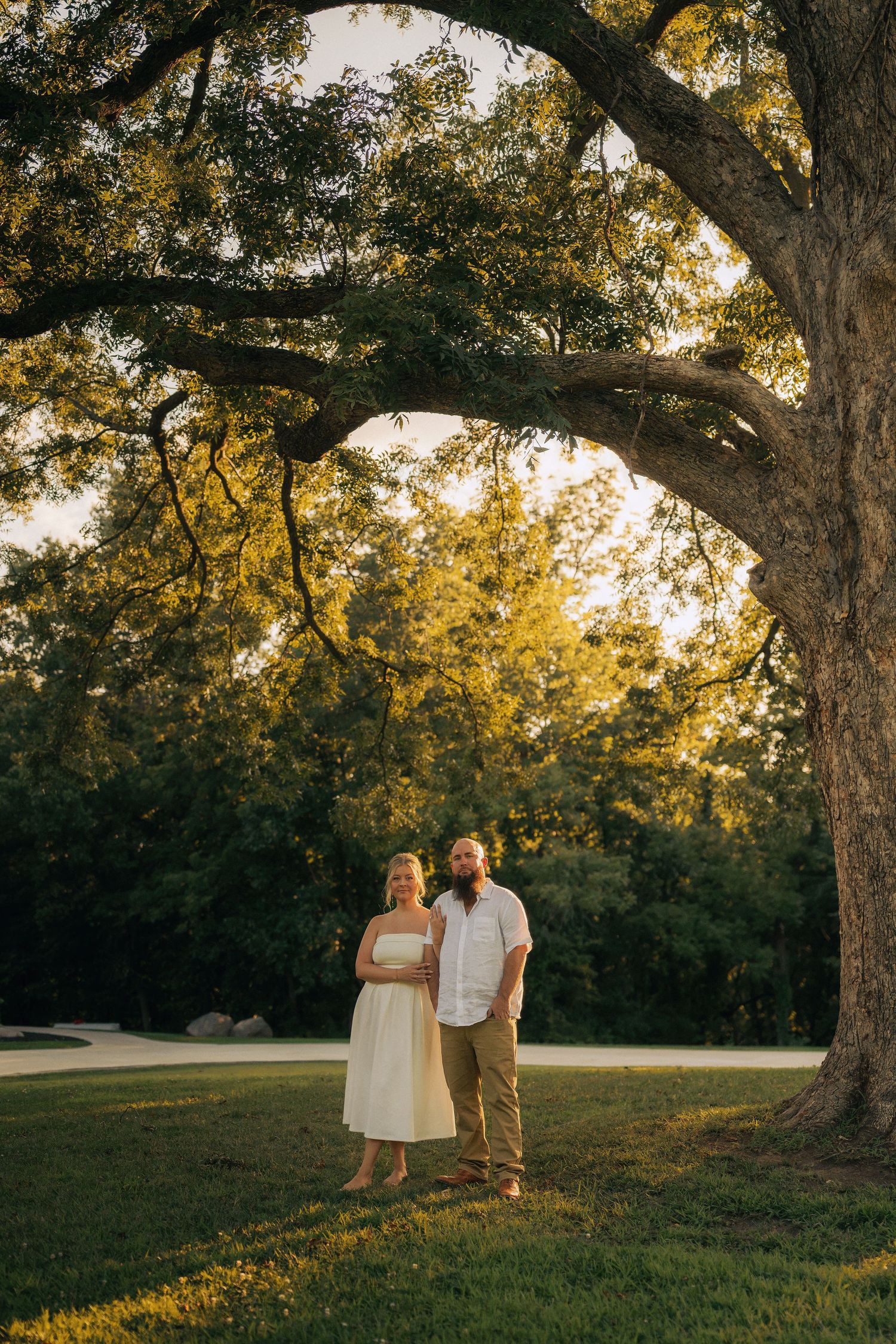 Couple standing under a large oak tree in golden hour sunlight on a green lawn.