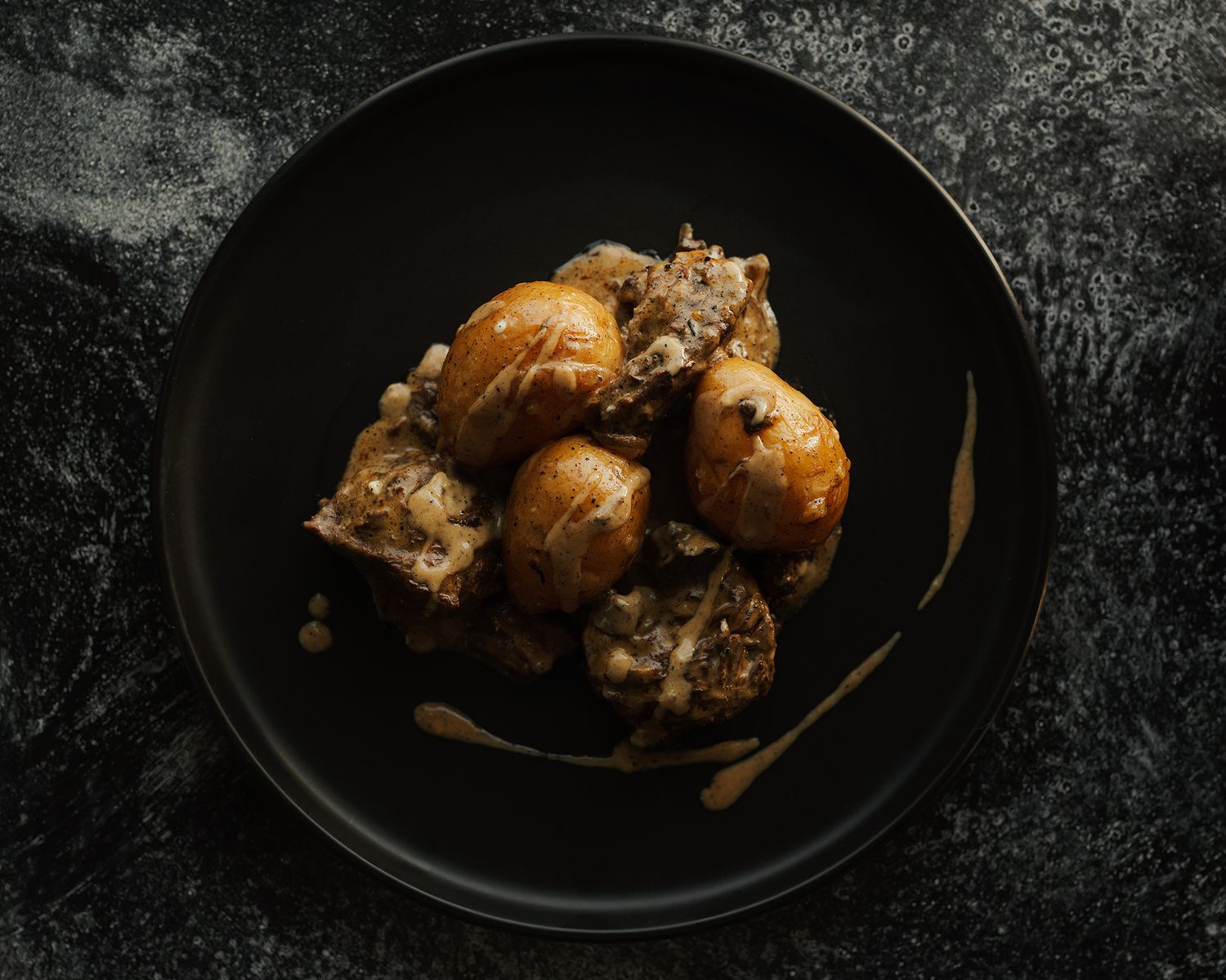 Roasted potatoes and braised meat served on a dark ceramic plate against a black textured background.