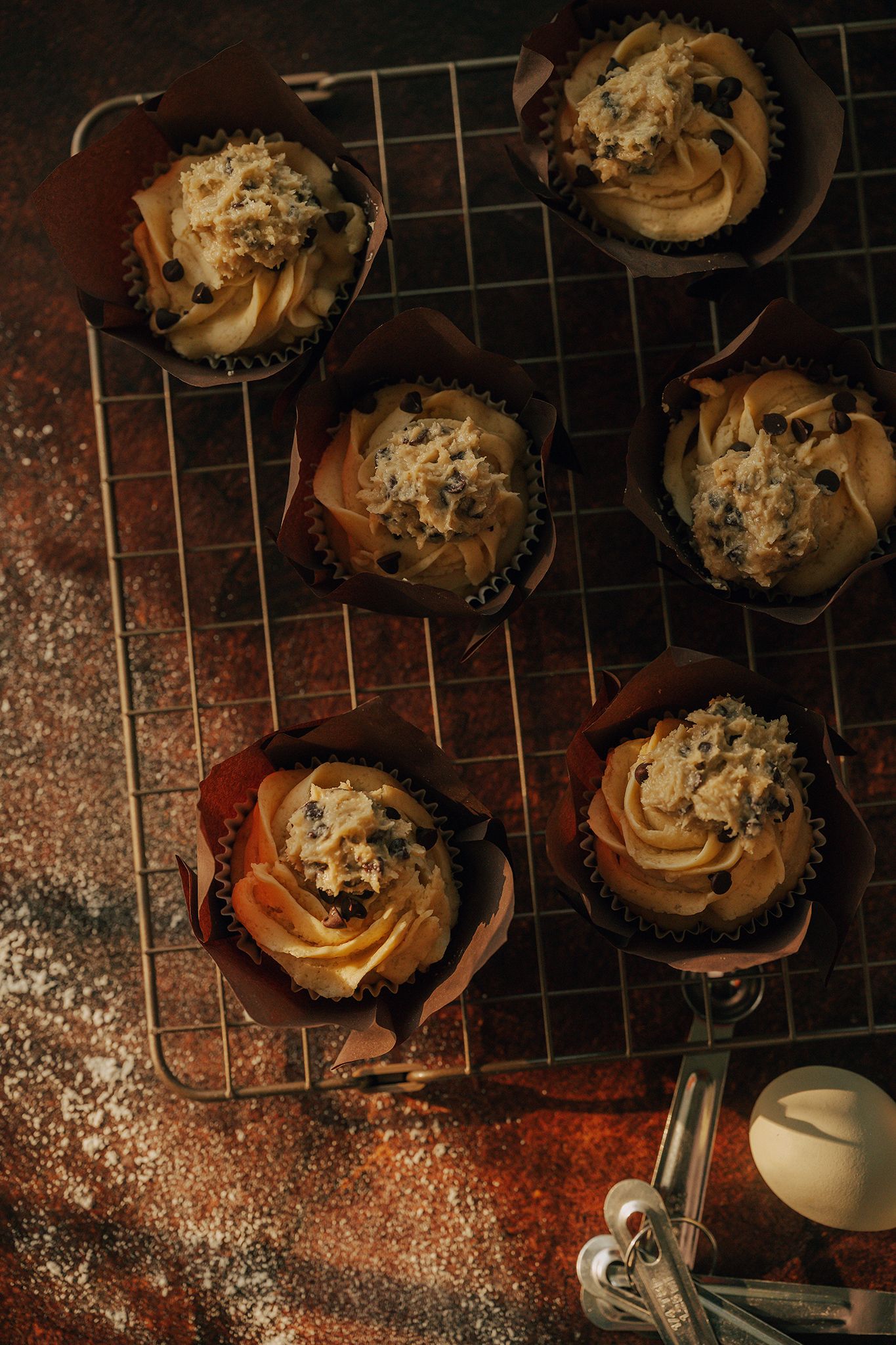 Six caramel frosted cupcakes with chocolate chips cooling on a wire rack on a dark rustic surface.