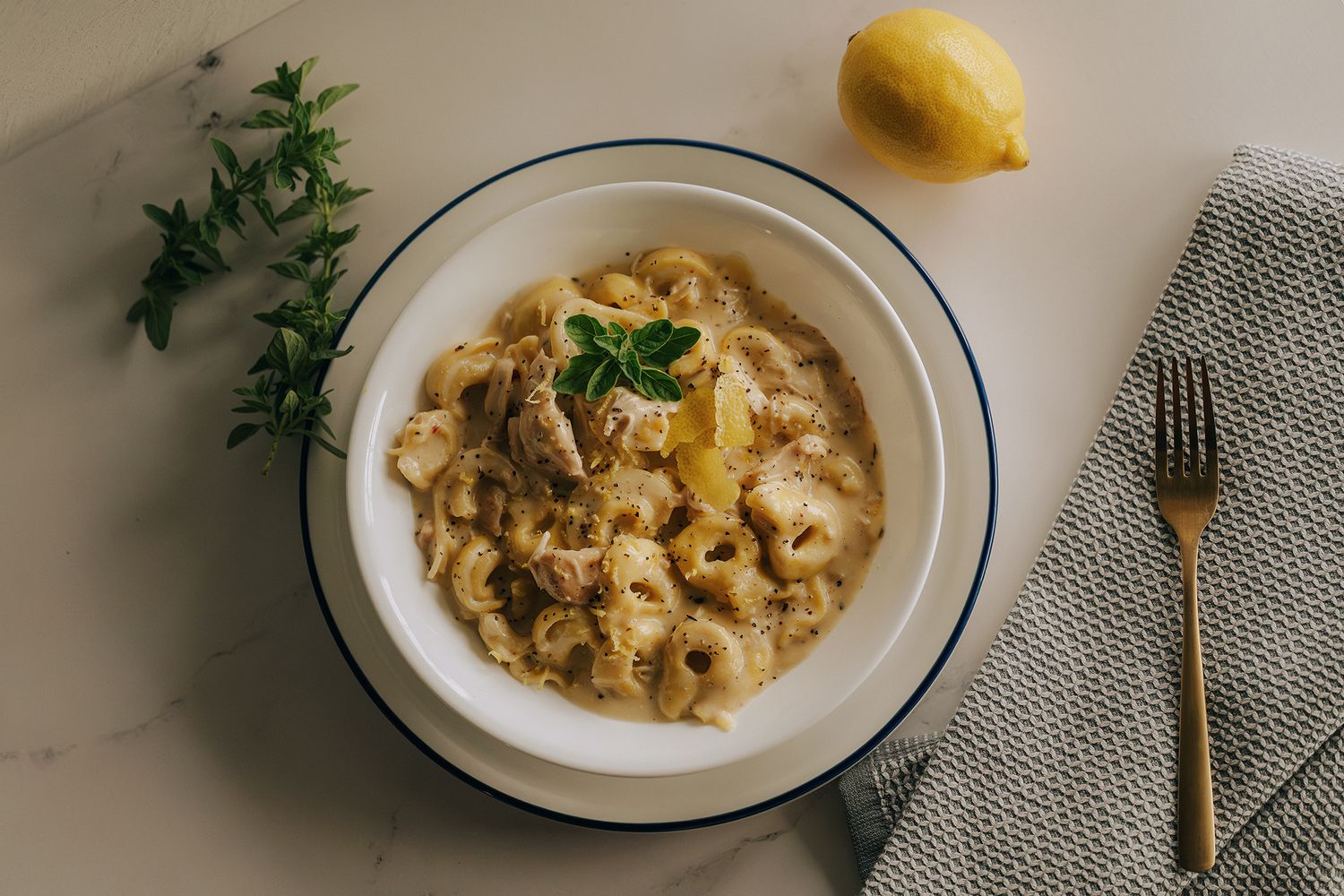 A bowl of creamy tortellini pasta garnished with fresh herbs, served with lemon on a marble surface.