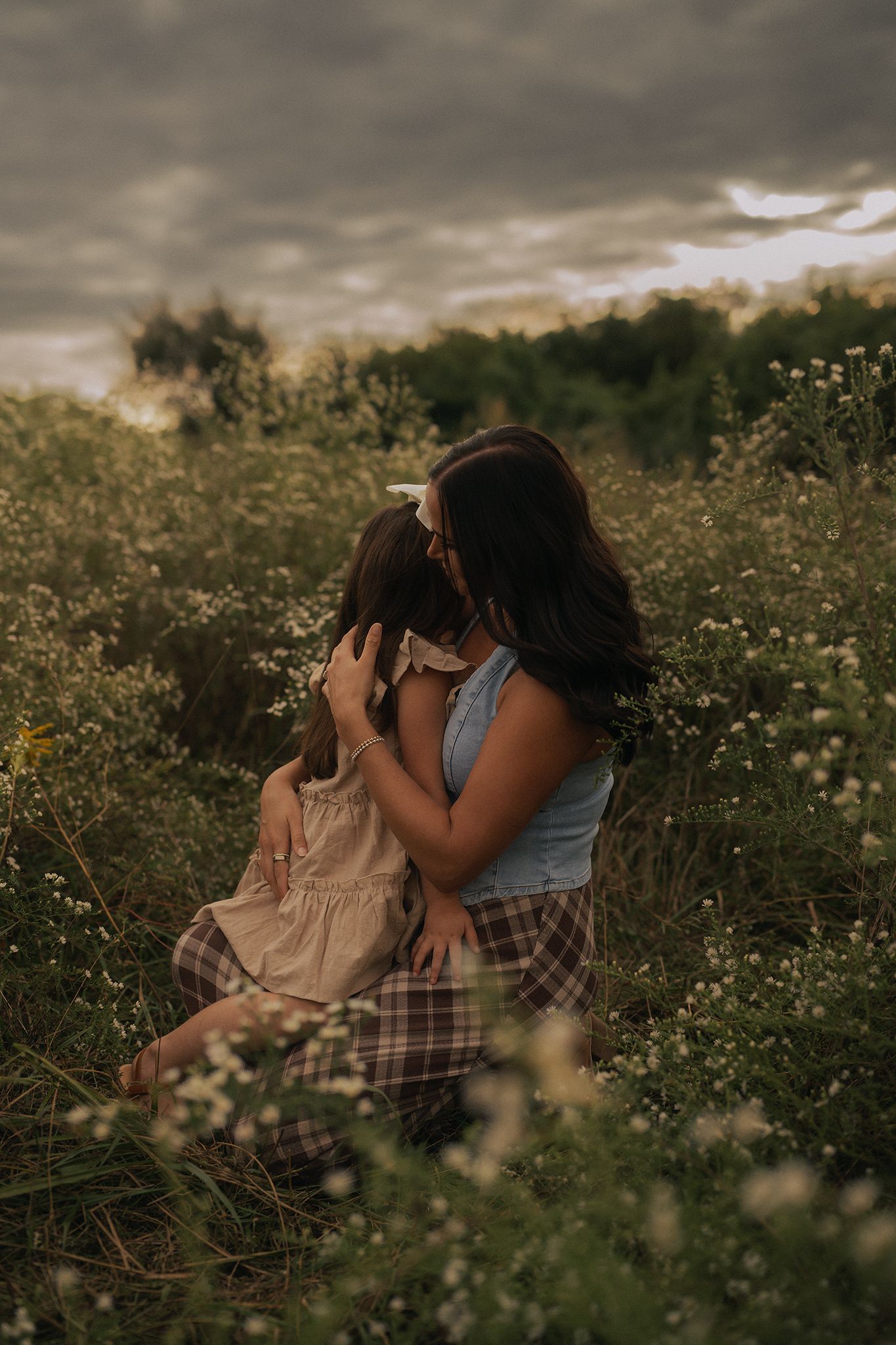 A mother and daughter share a tender embrace in a wildflower field under a dramatic cloudy sky.