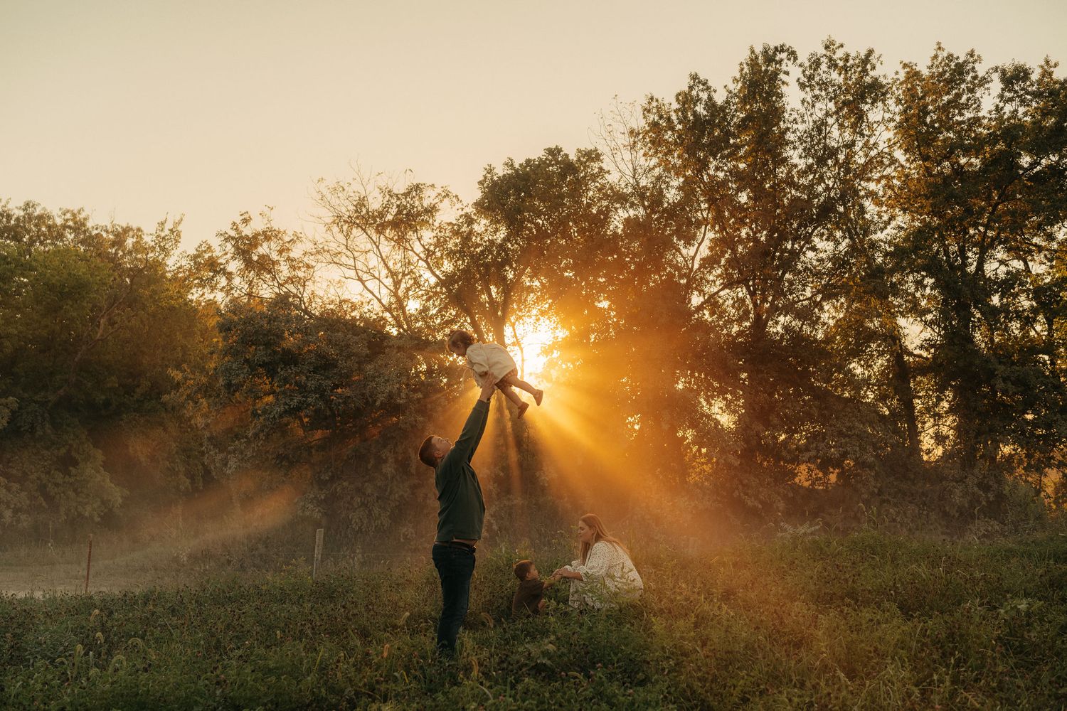 A person lifts a child toward golden rays of sunlight streaming through misty trees at sunset.