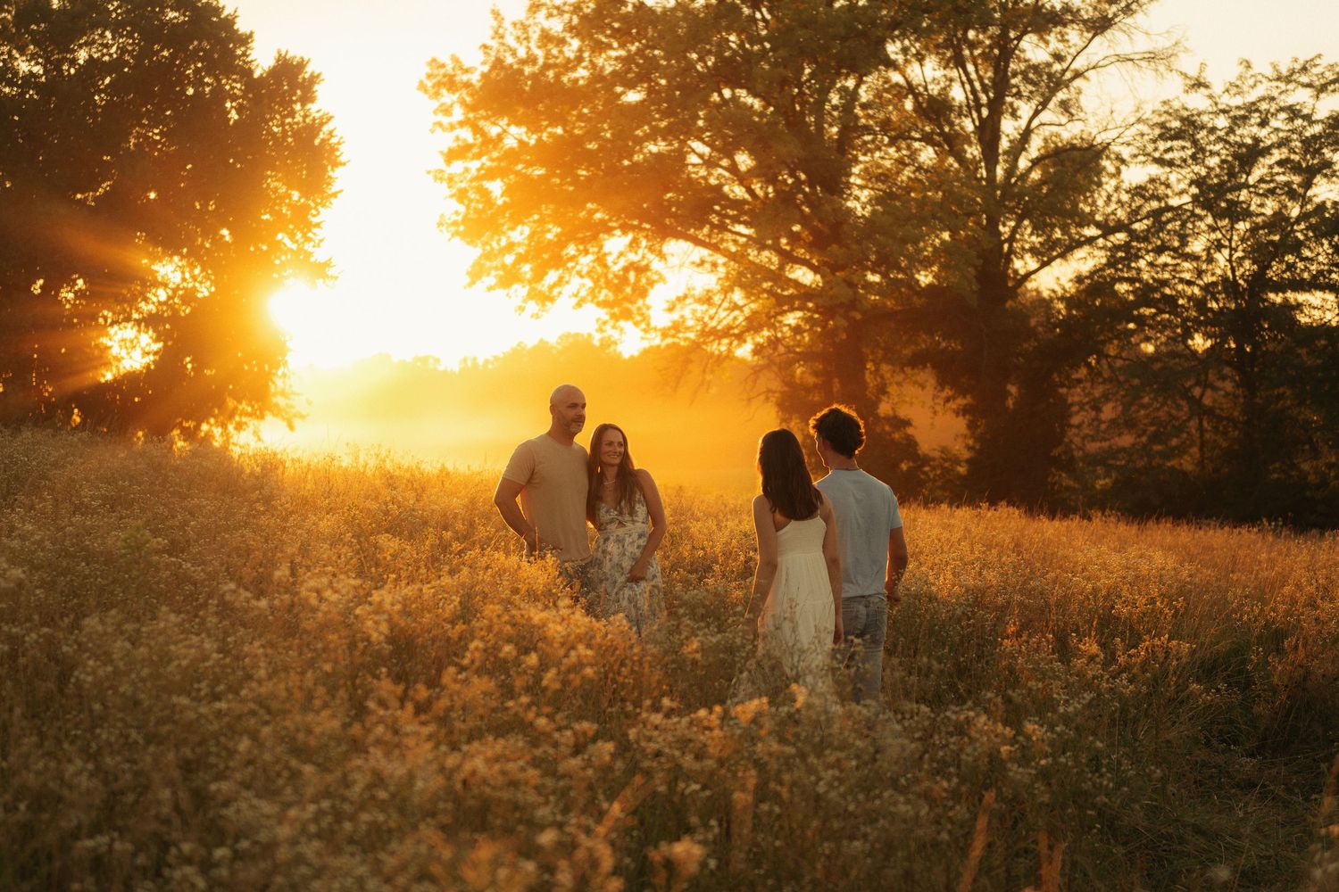 Family of four standing in golden meadow at sunset, surrounded by wildflowers and glowing warm light.