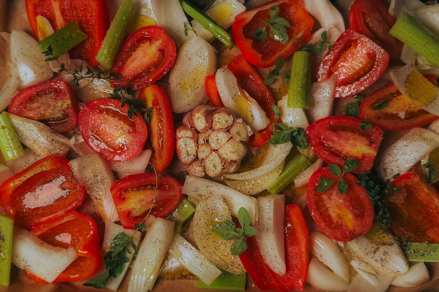 Fresh mixed vegetables including halved tomatoes, celery, red peppers, onions, and garlic cloves ready for roasting.