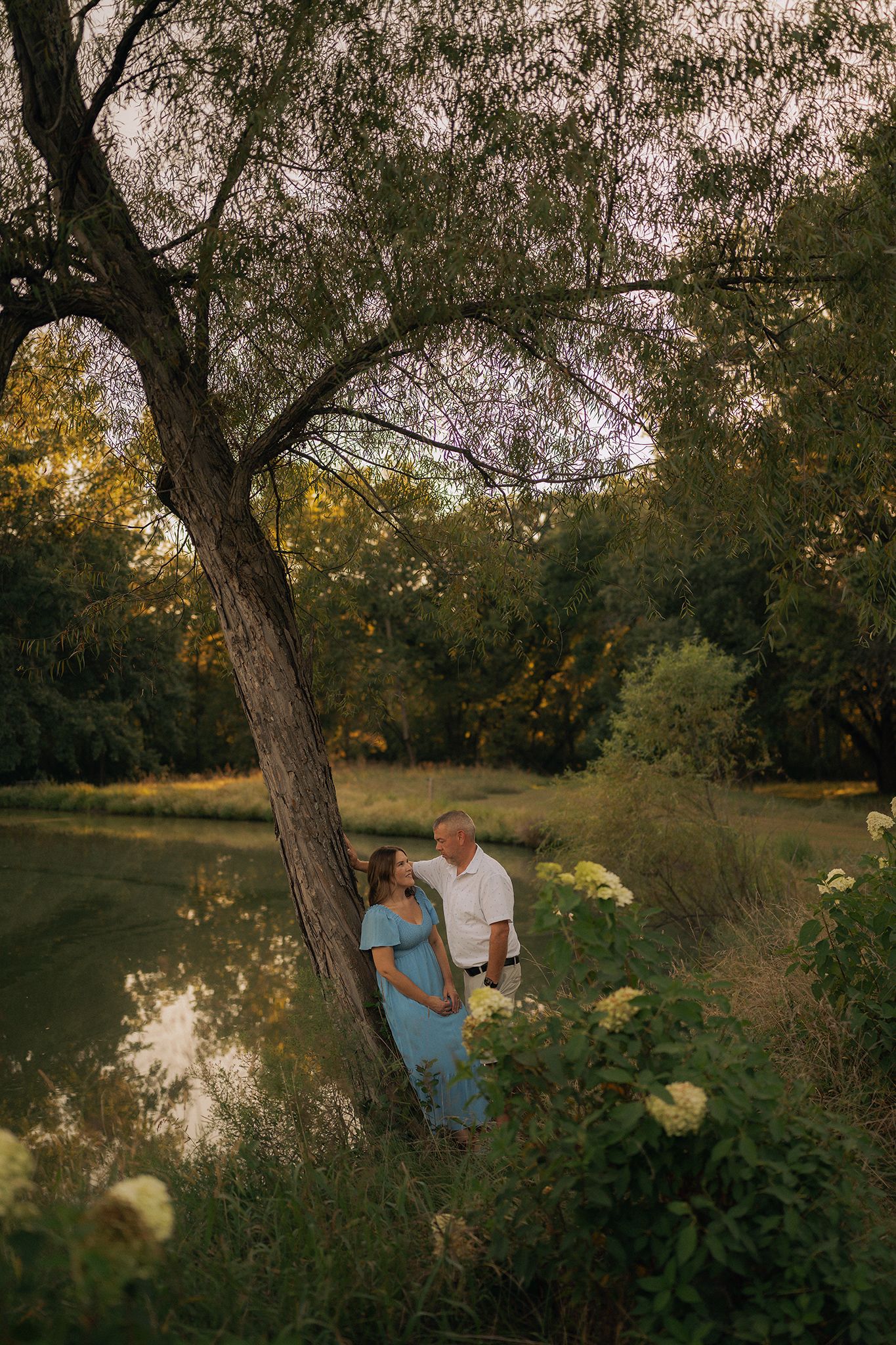 Couple standing under a large tree by a pond in a lush green park during golden hour maternity photo session.