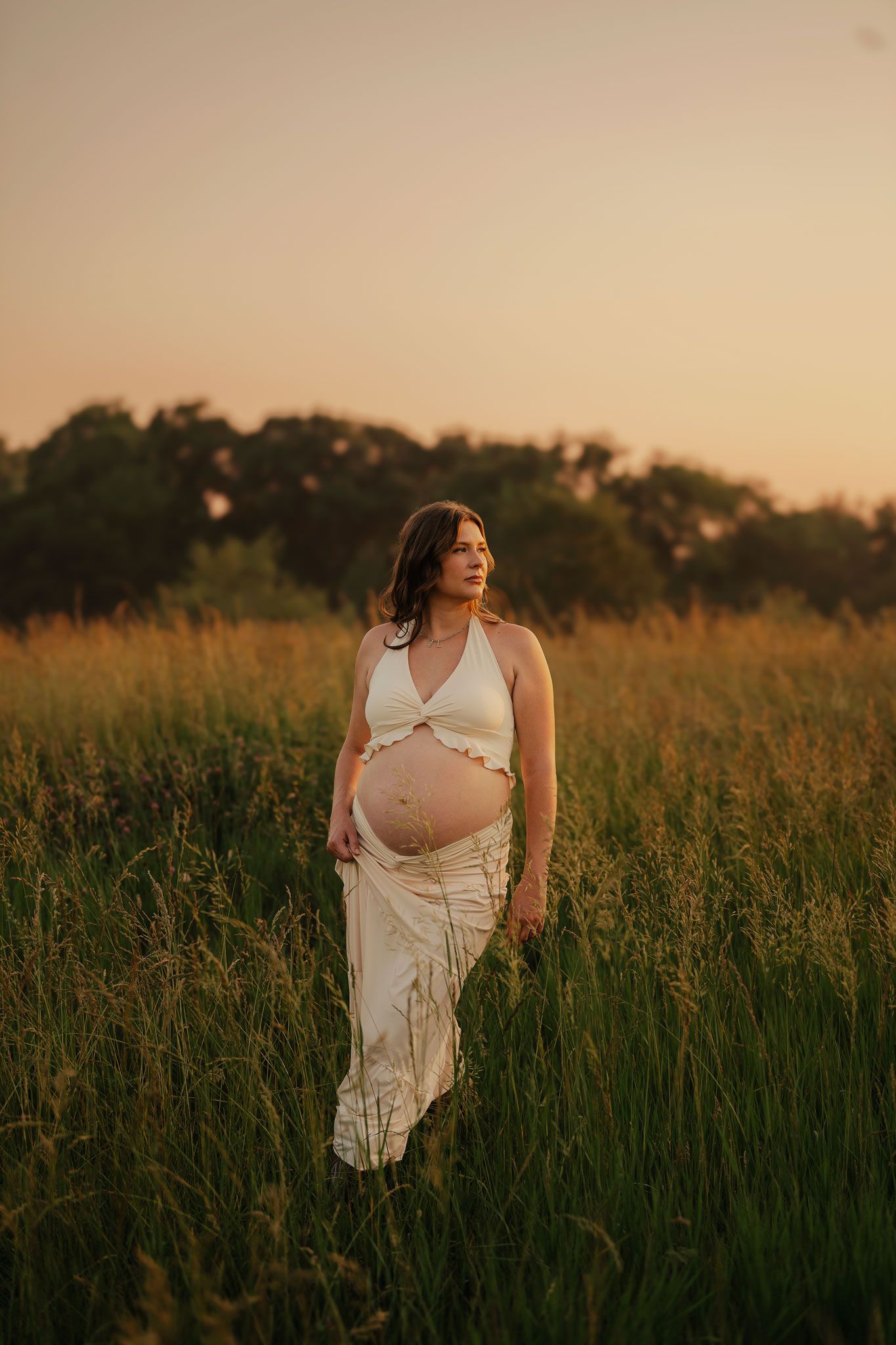 Pregnant woman in white crop top and skirt standing in golden field at sunset.