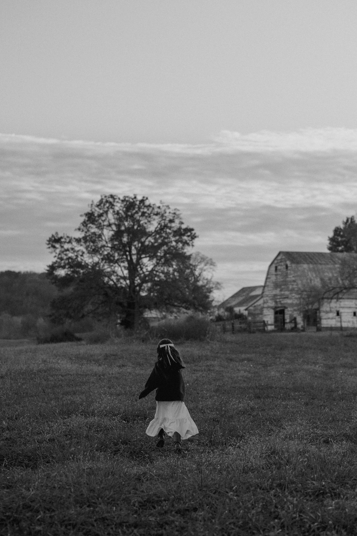 Black and white photo of a young girl walking toward a rustic barn and tree in an open field at dusk.