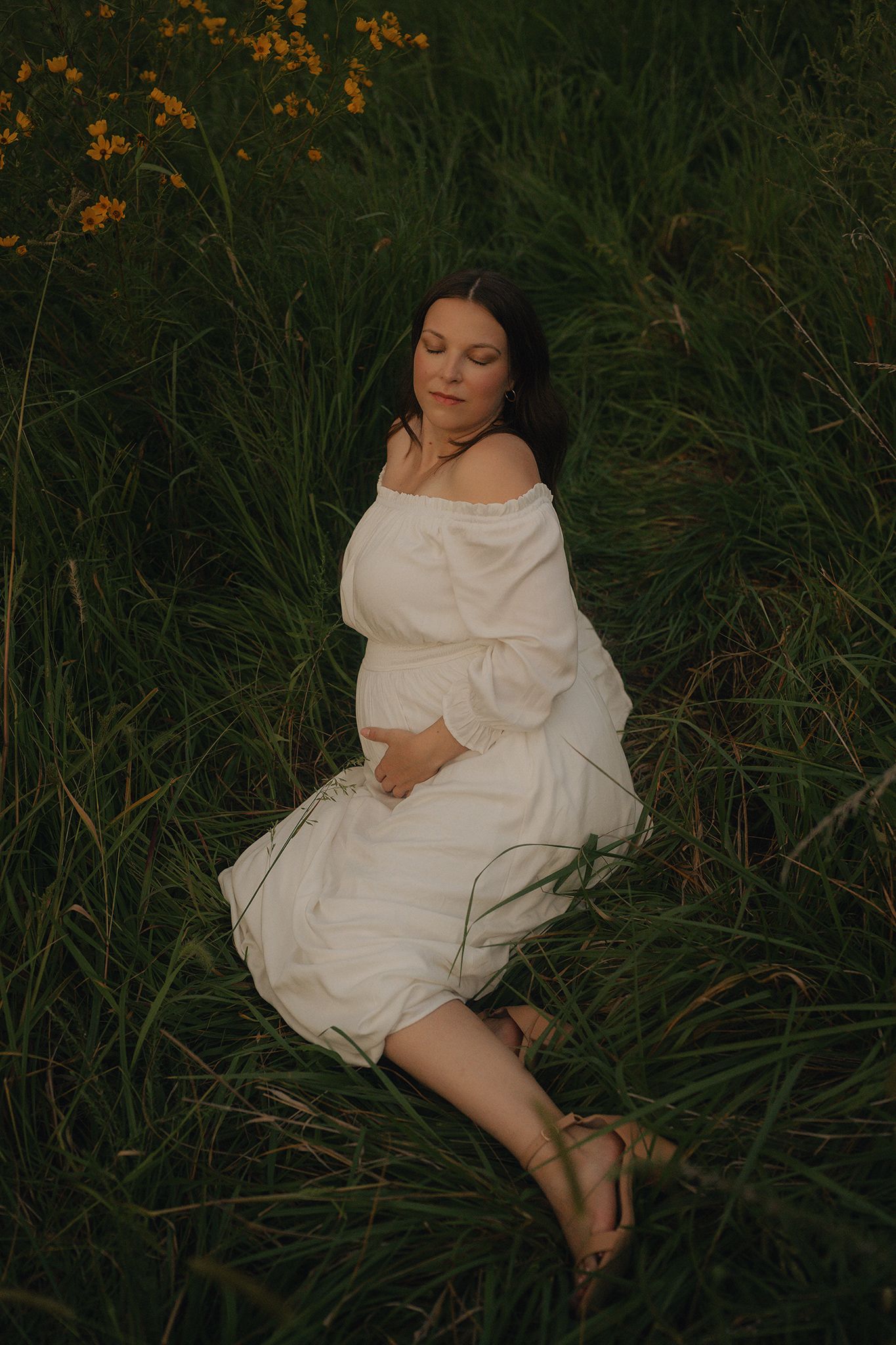 Pregnant woman in white off-shoulder dress sitting peacefully in tall green grass with yellow wildflowers.