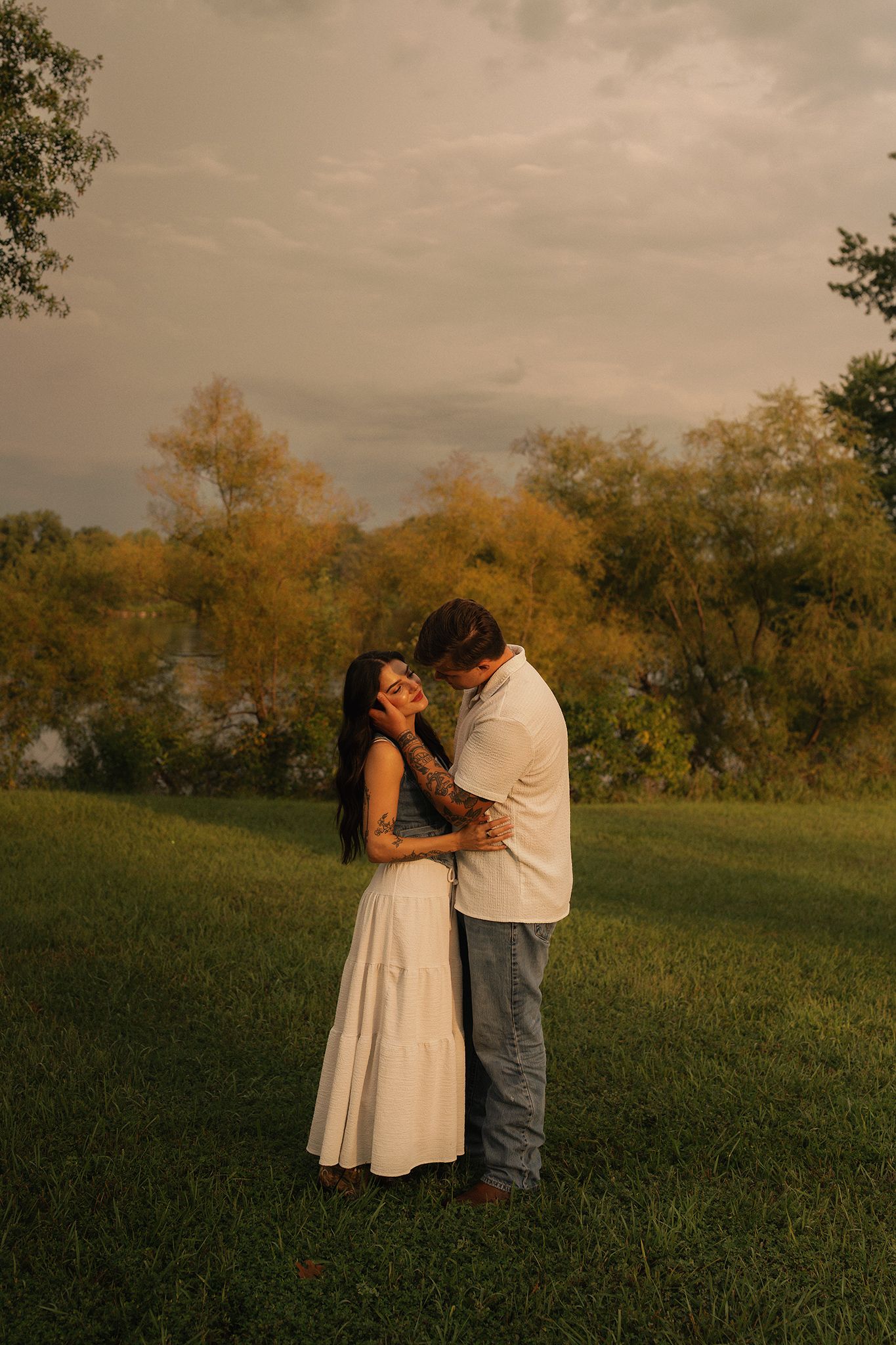 Couple embracing in a green field at golden hour, woman in white dress, trees with autumn foliage in background.