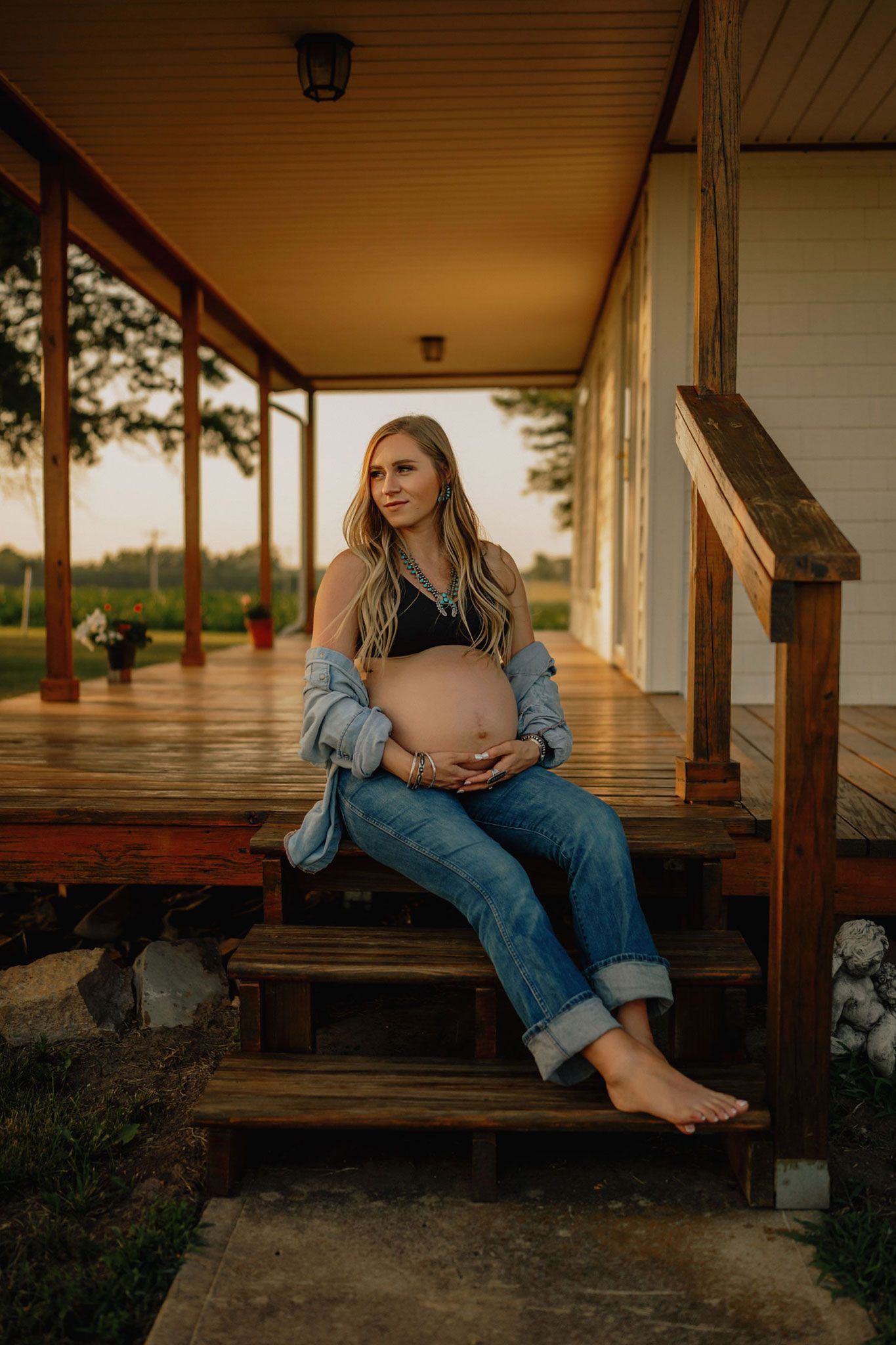Pregnant woman sitting on wooden porch steps at sunset, cradling baby bump, wearing jeans and denim shirt.