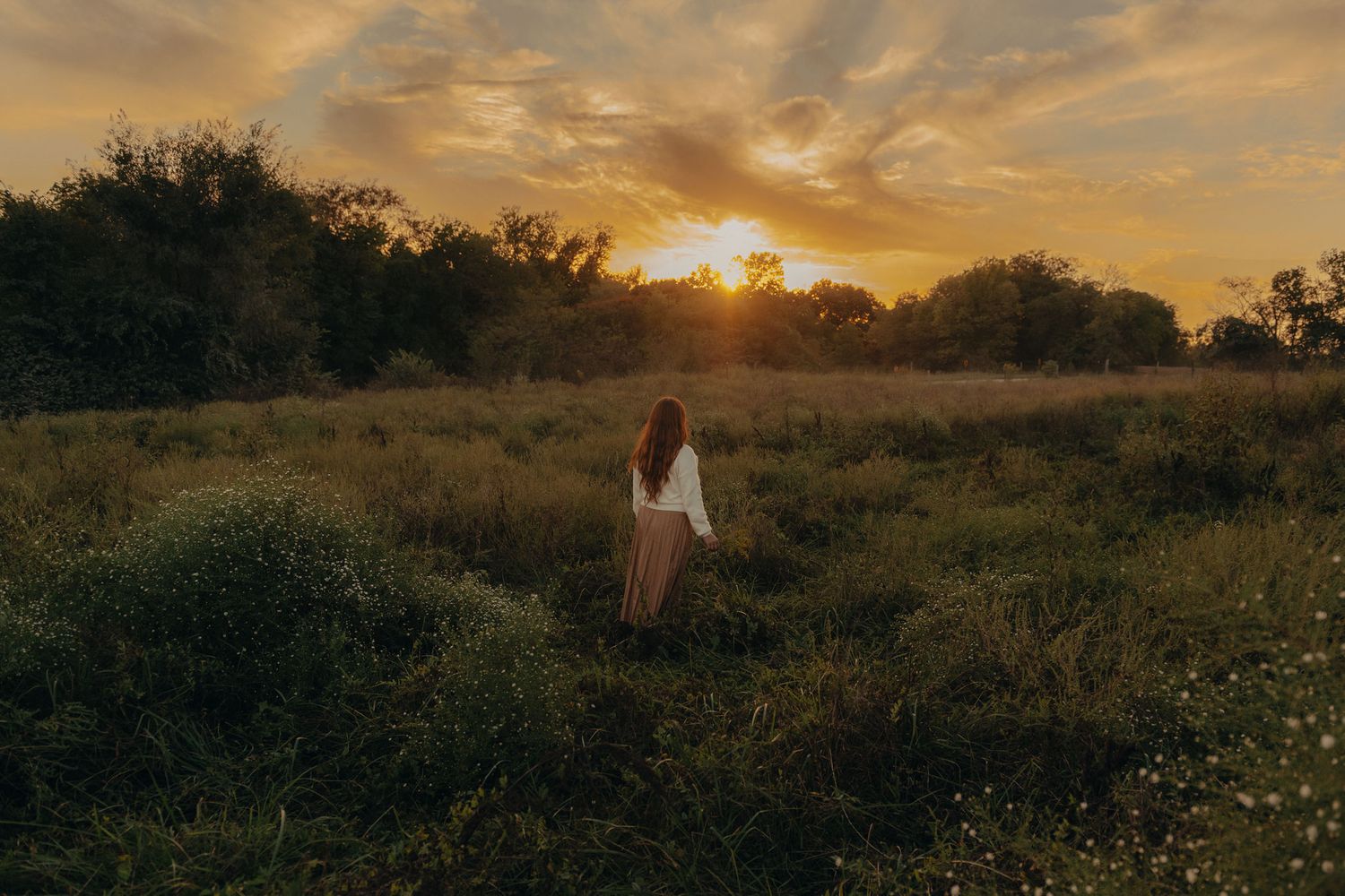 A woman in white stands alone in a lush meadow facing a dramatic golden sunset sky.