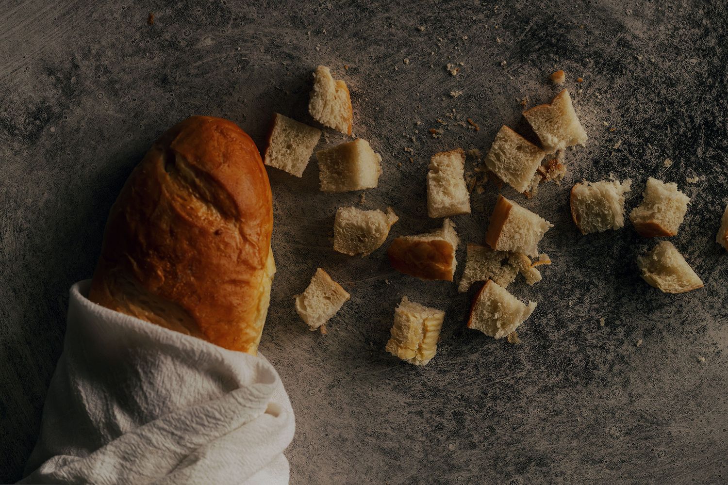 A rustic bread loaf wrapped in white linen beside scattered bread cubes on a dark stone surface.