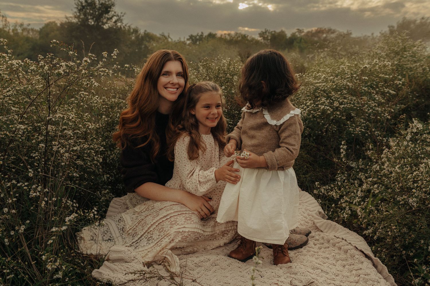 A mother with red hair sits with two young daughters on a blanket among wildflowers.