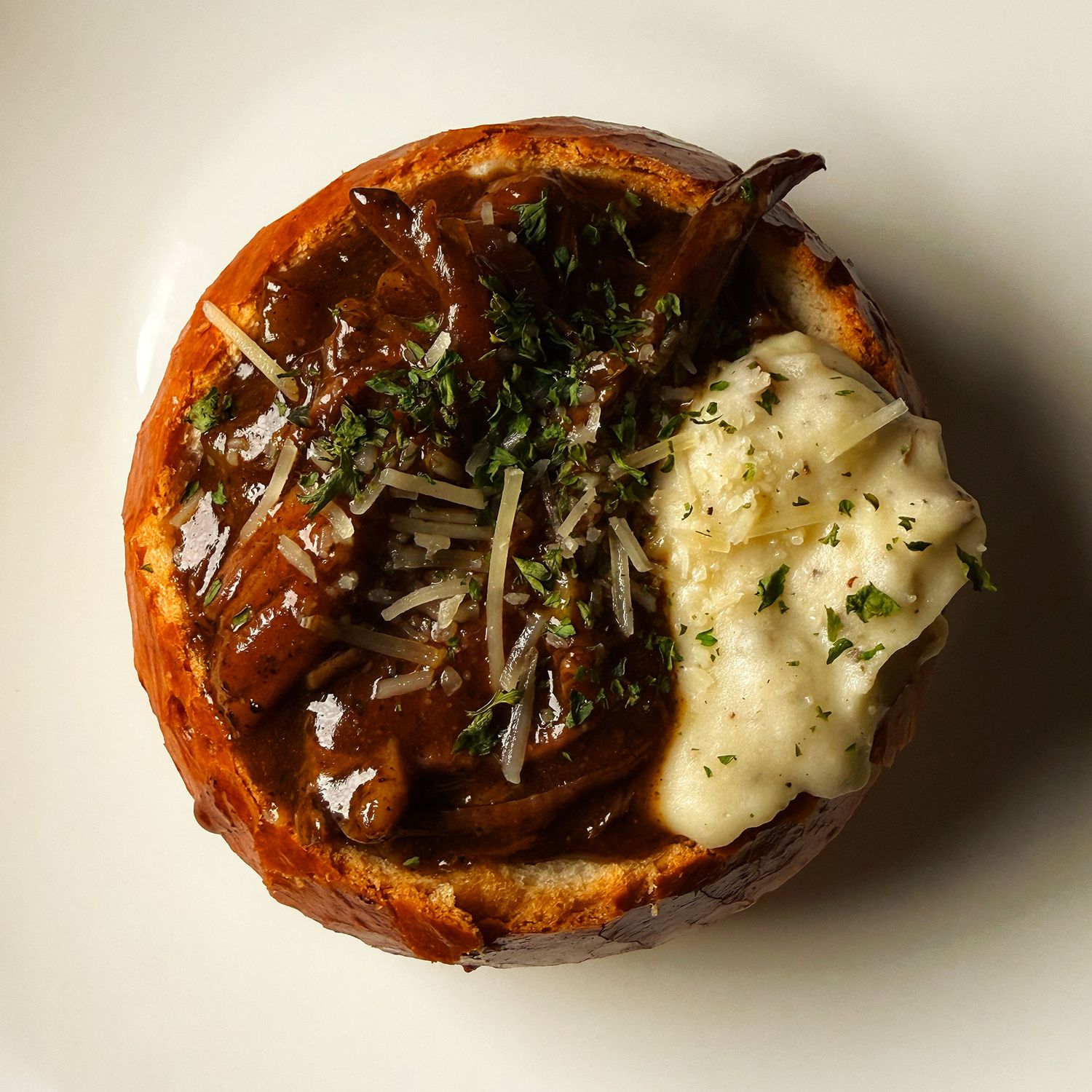 French onion roast soup in a bread bowl topped with melted cheese, caramelized onions, and fresh herbs on a white plate.