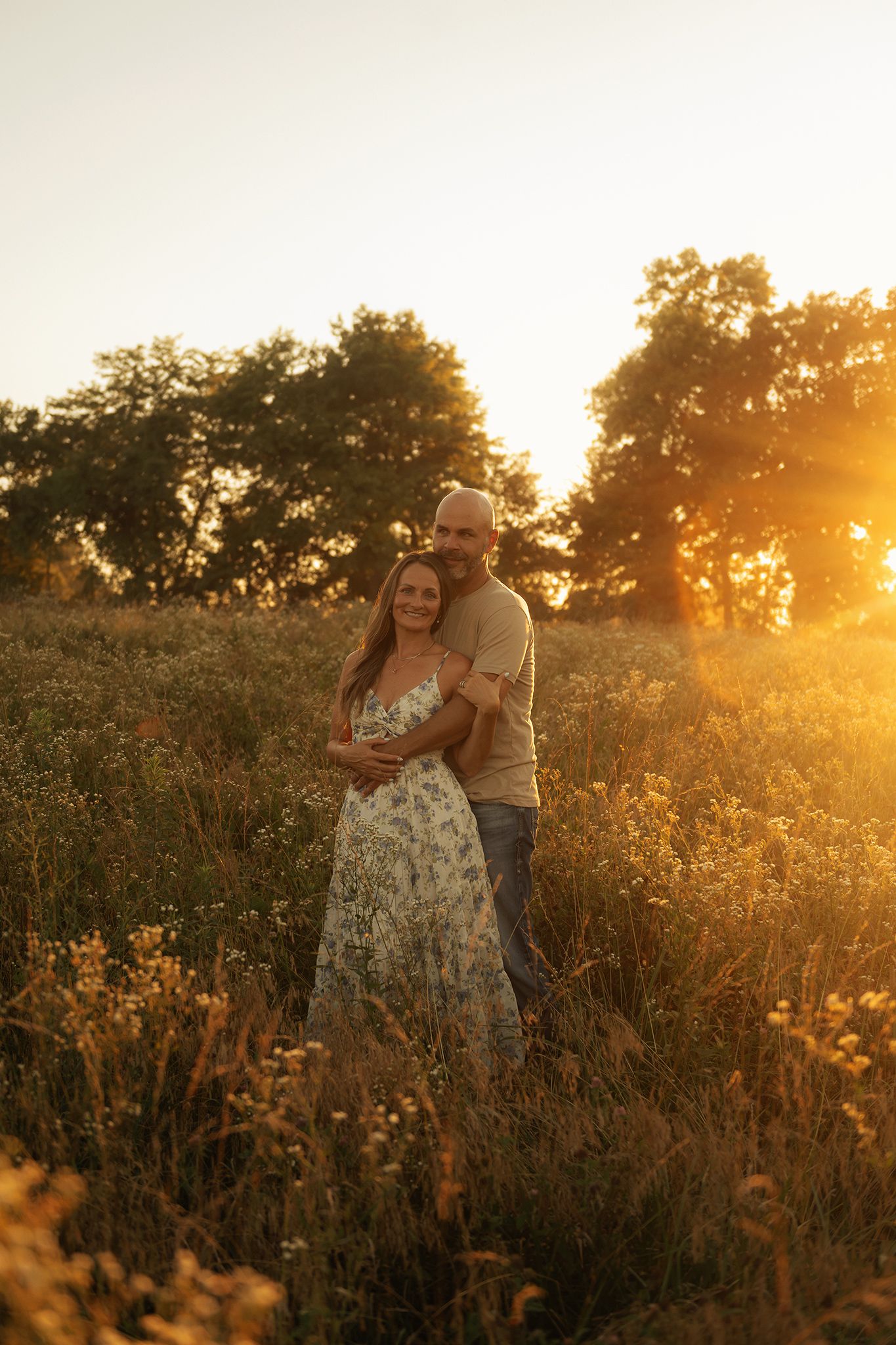Couple embracing in a golden sunlit meadow with wildflowers and trees at sunset.