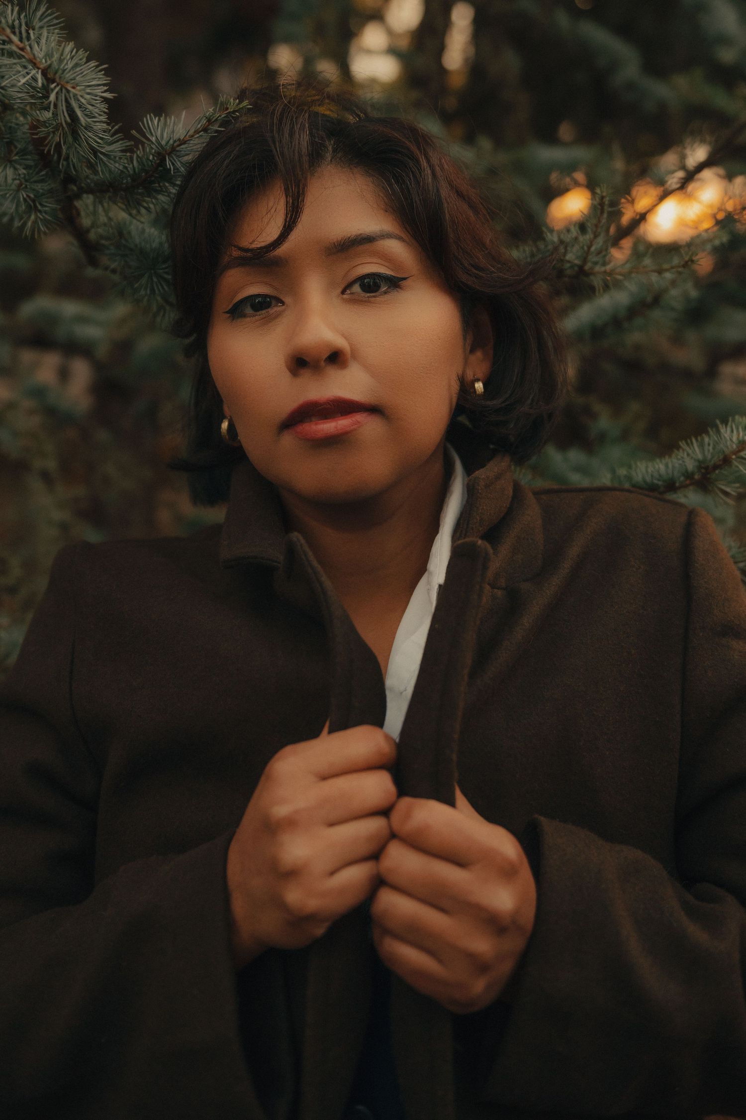 A young woman in a dark coat holds her lapels while standing among evergreen trees in soft golden light.