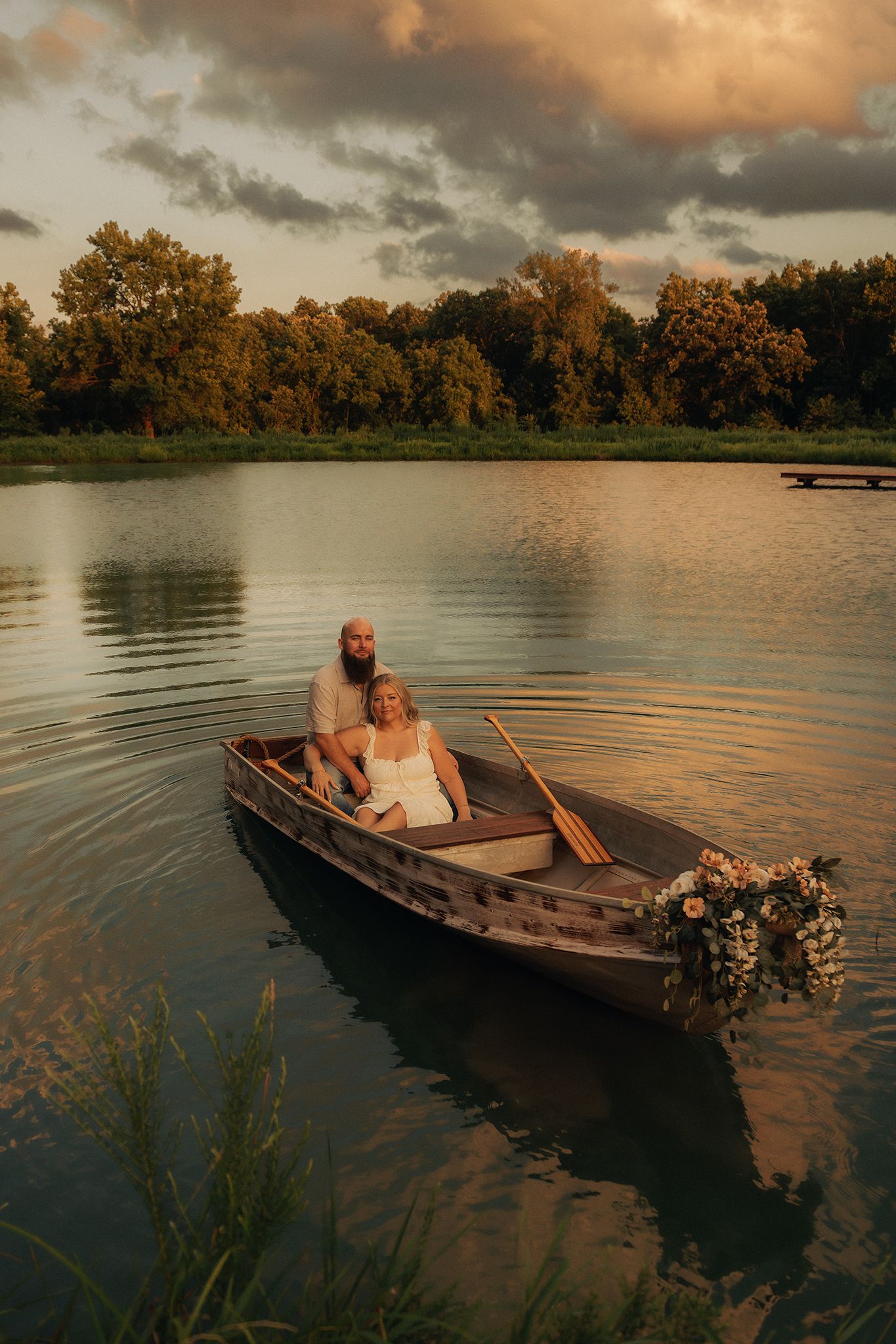 Couple sitting in a rustic rowboat with flowers at sunset on a calm lake surrounded by lush green trees.