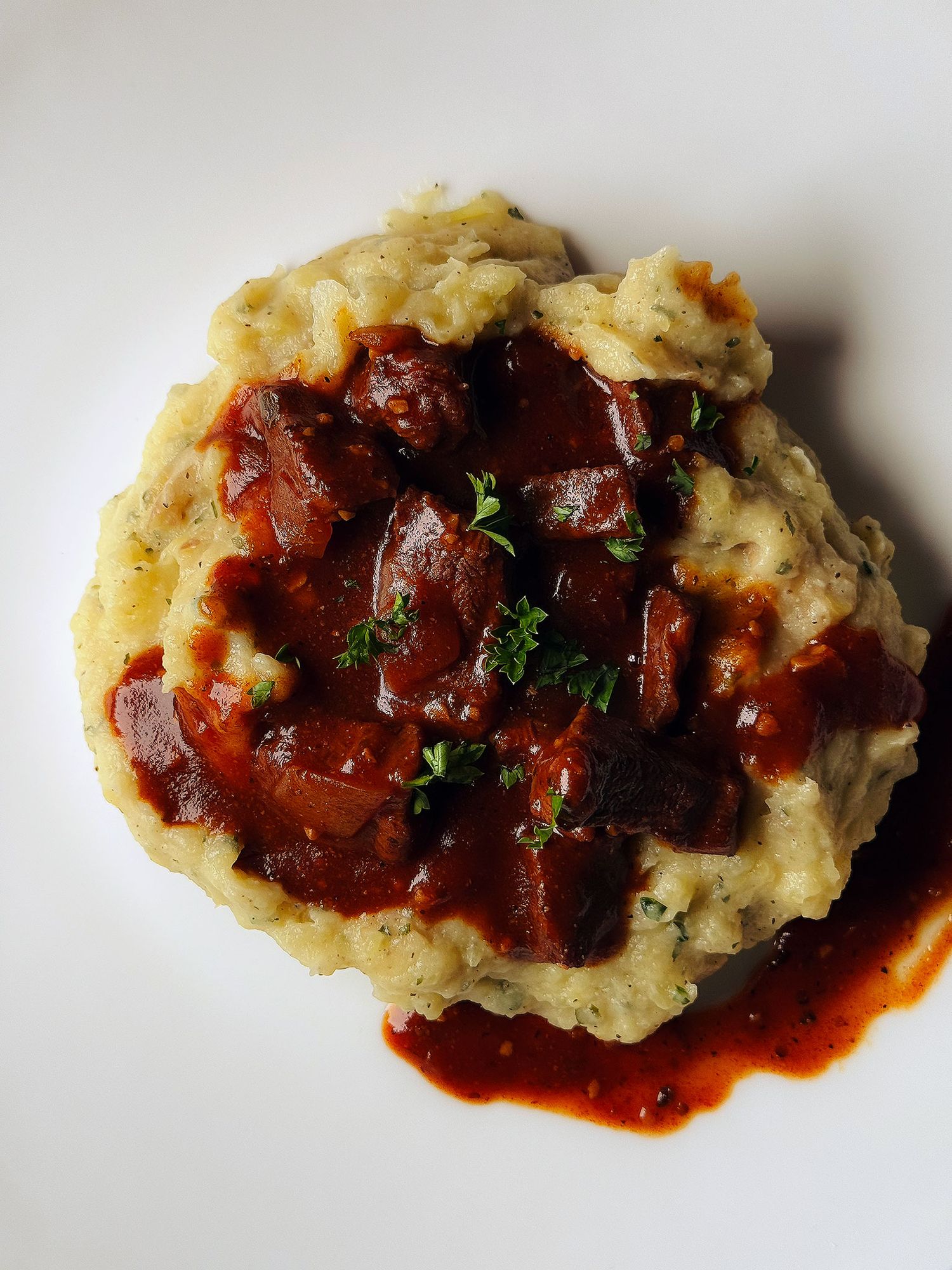 Mashed potatoes topped with rich tomato braised meat chunks and fresh herbs on a white plate.