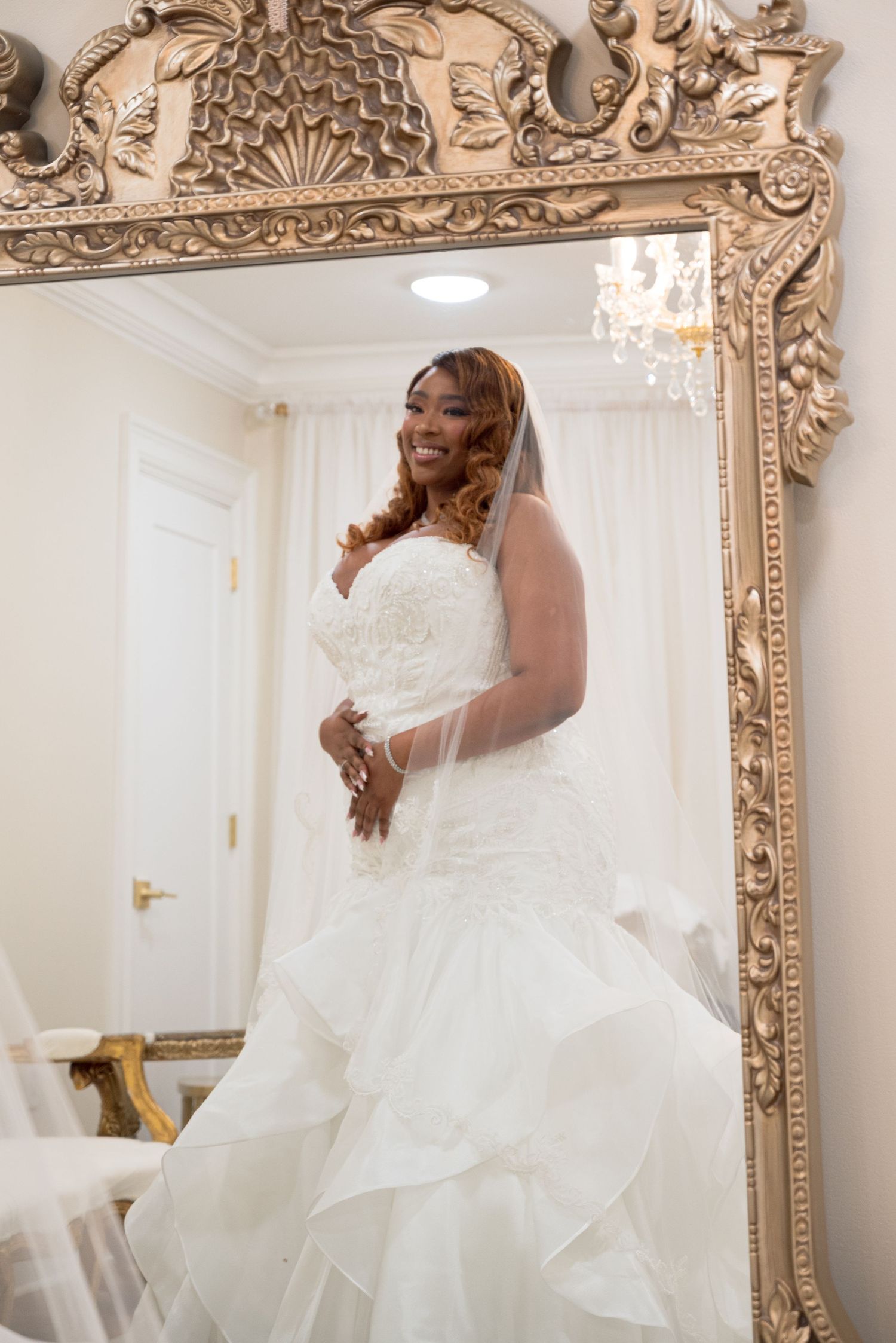 Elegant bride in lace mermaid wedding gown poses before a large ornate gold-framed mirror.
